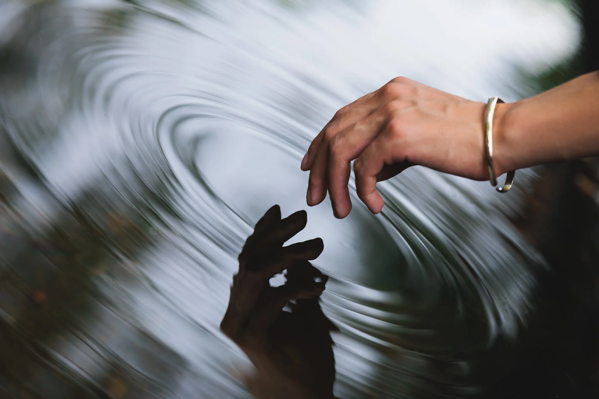 A person's hand touching a reflective water surface with ripples, wearing a silver bracelet.