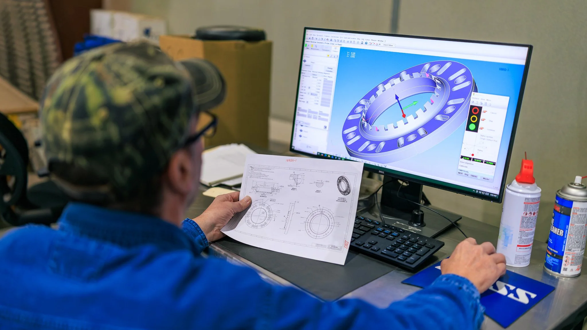 A person wearing a blue jacket and camouflage helmet examining technical engineering drawings at a workstation, with a computer monitor displaying a 3D model of a mechanical part.