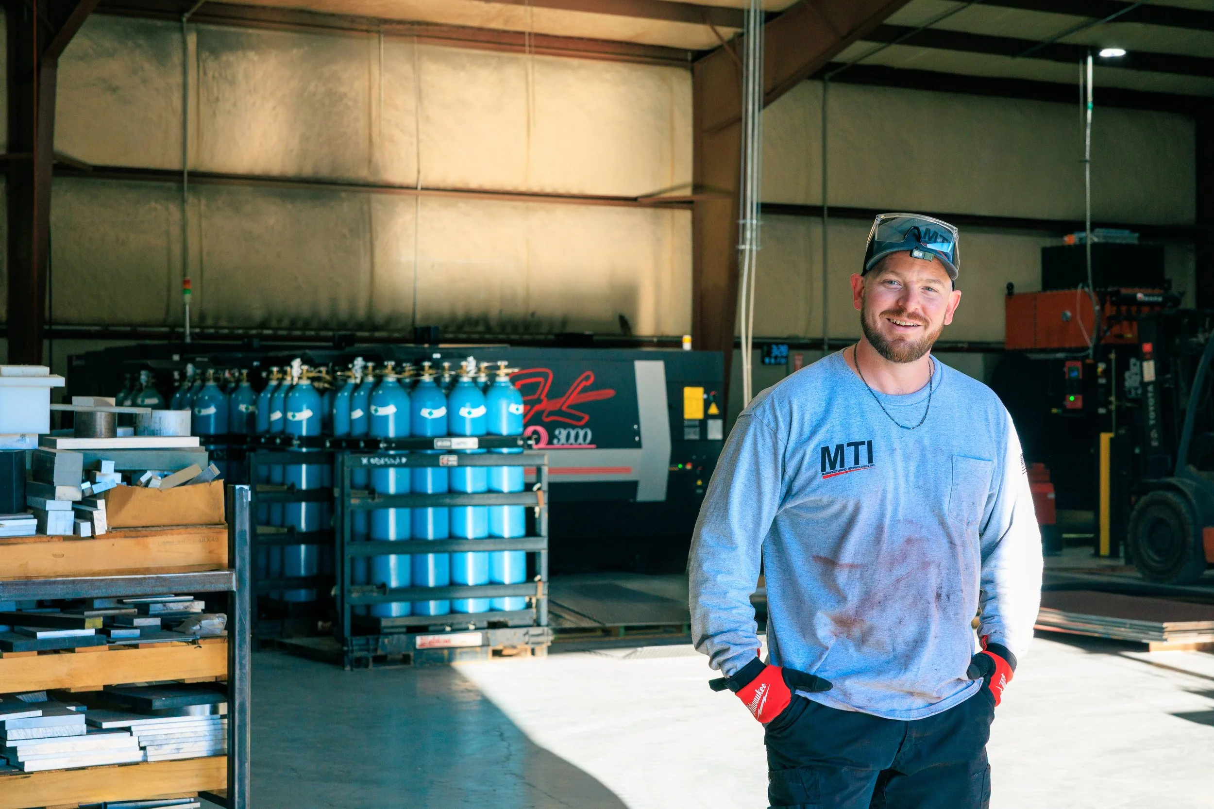A smiling man wearing a cap, gray sweatshirt, safety gloves, and work pants standing in an industrial workshop or warehouse.