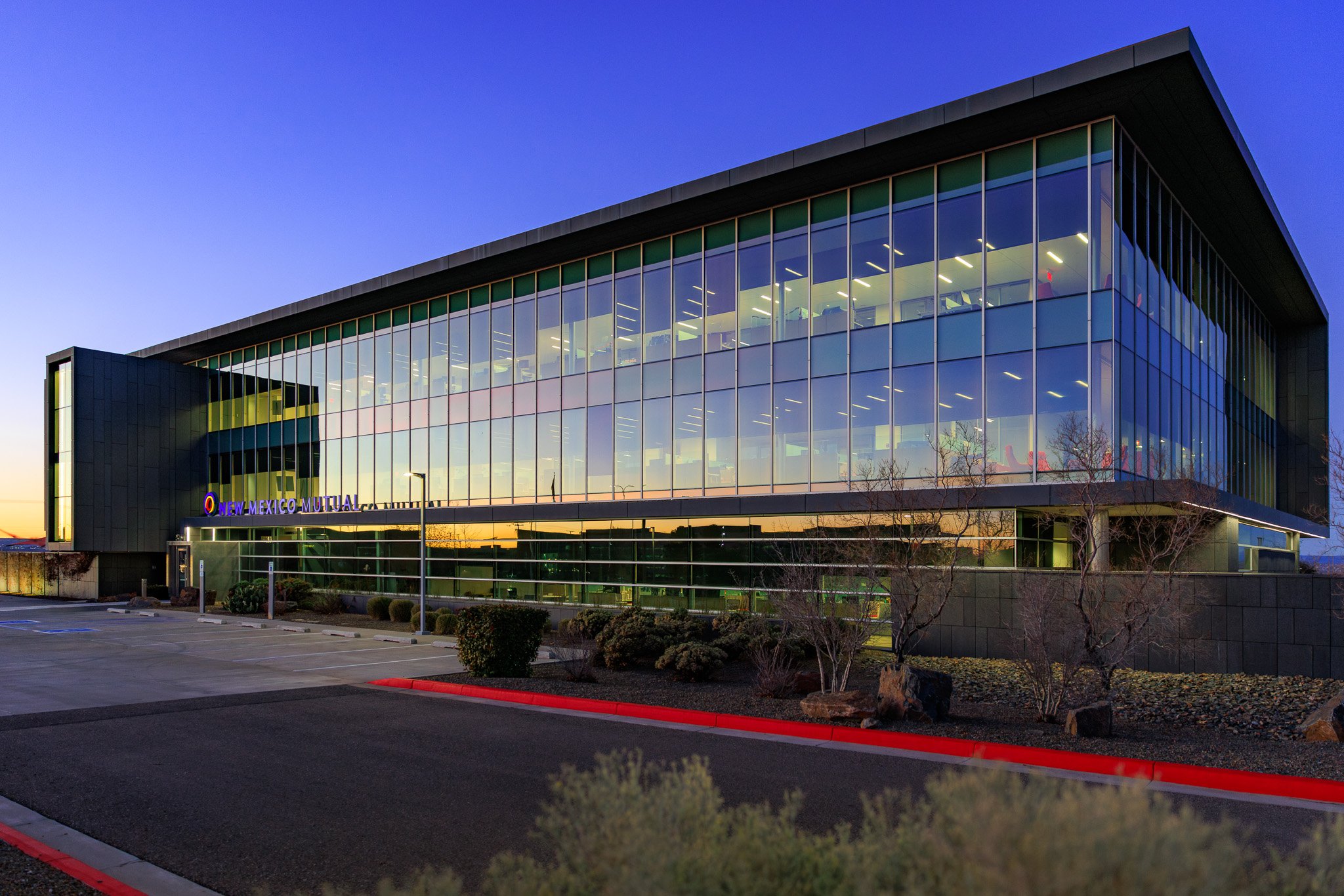 Modern glass building with illuminated interior, indicating it is evening, with a parking lot in the foreground and trees and shrubs around.