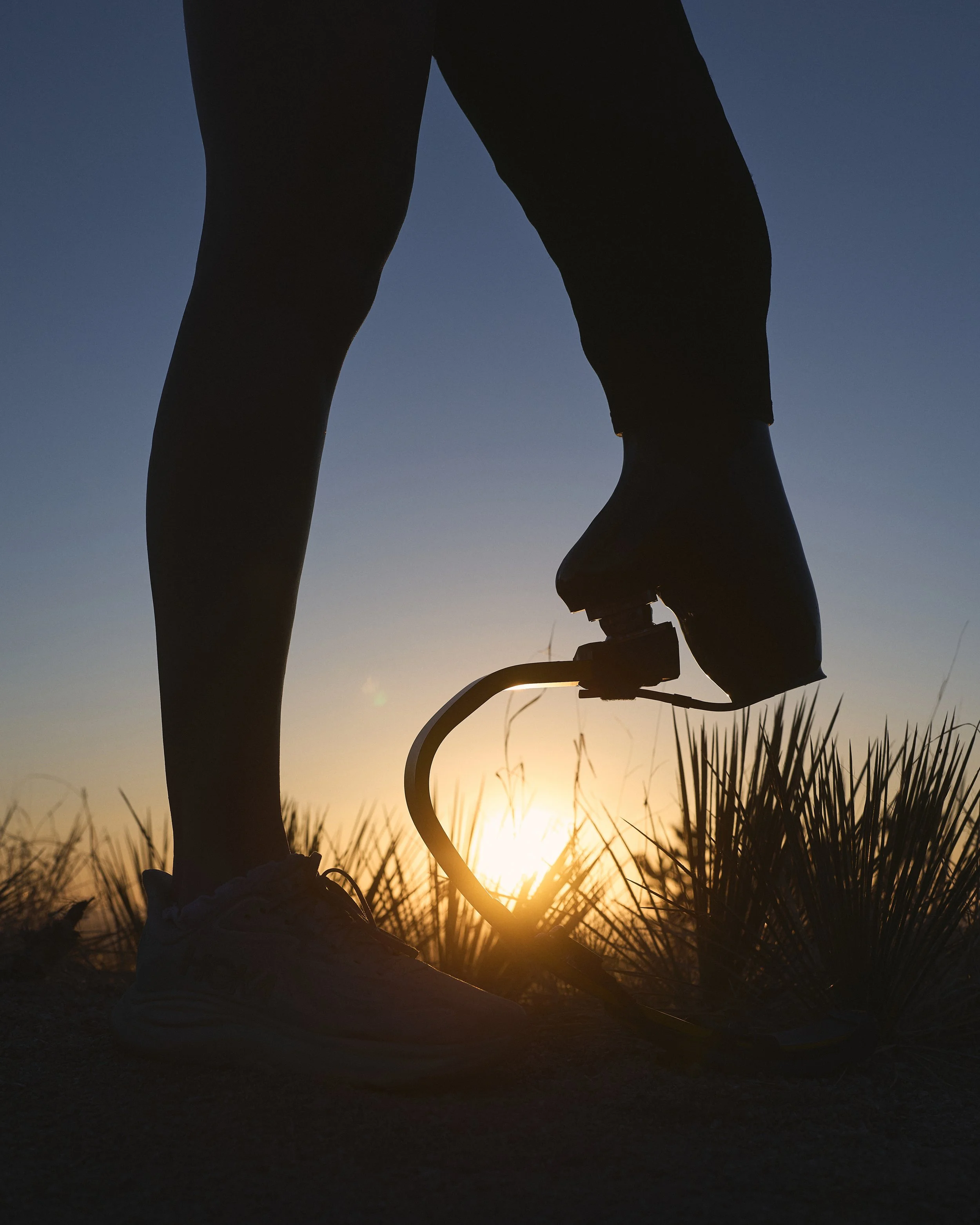 Silhouette of a person standing next to a bicycle on a grassy hill at sunset.