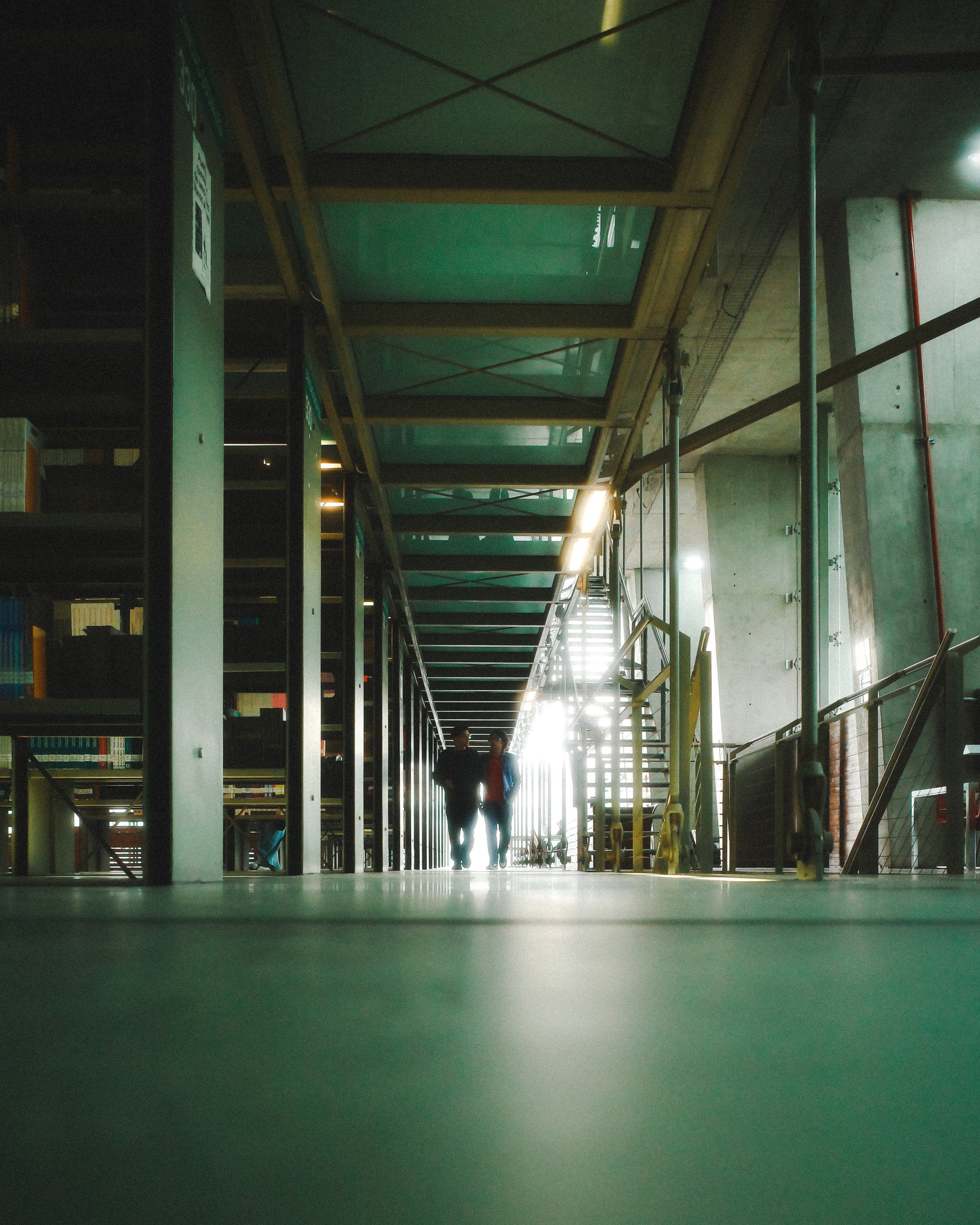 Two people walking down a corridor in a modern building, backlit by sunlight streaming through a glass door at the end of the corridor. There are bookshelves on the left side of the corridor, and construction scaffolding on the right side.