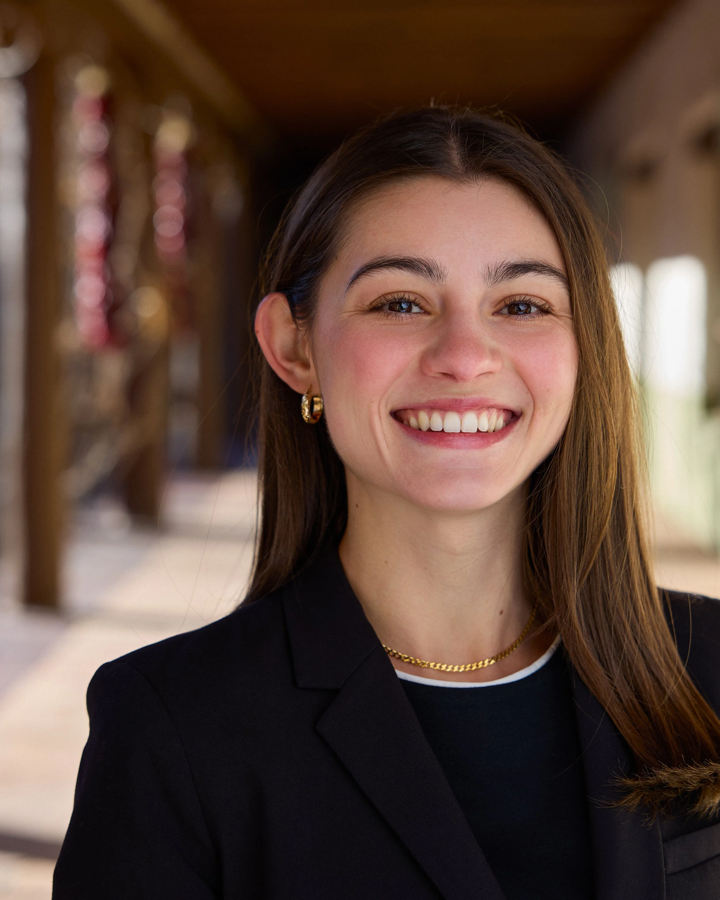 Close-up of a smiling woman with long brown hair, wearing gold hoop earrings, a gold chain necklace, a black blazer, and a white shirt with black trim.