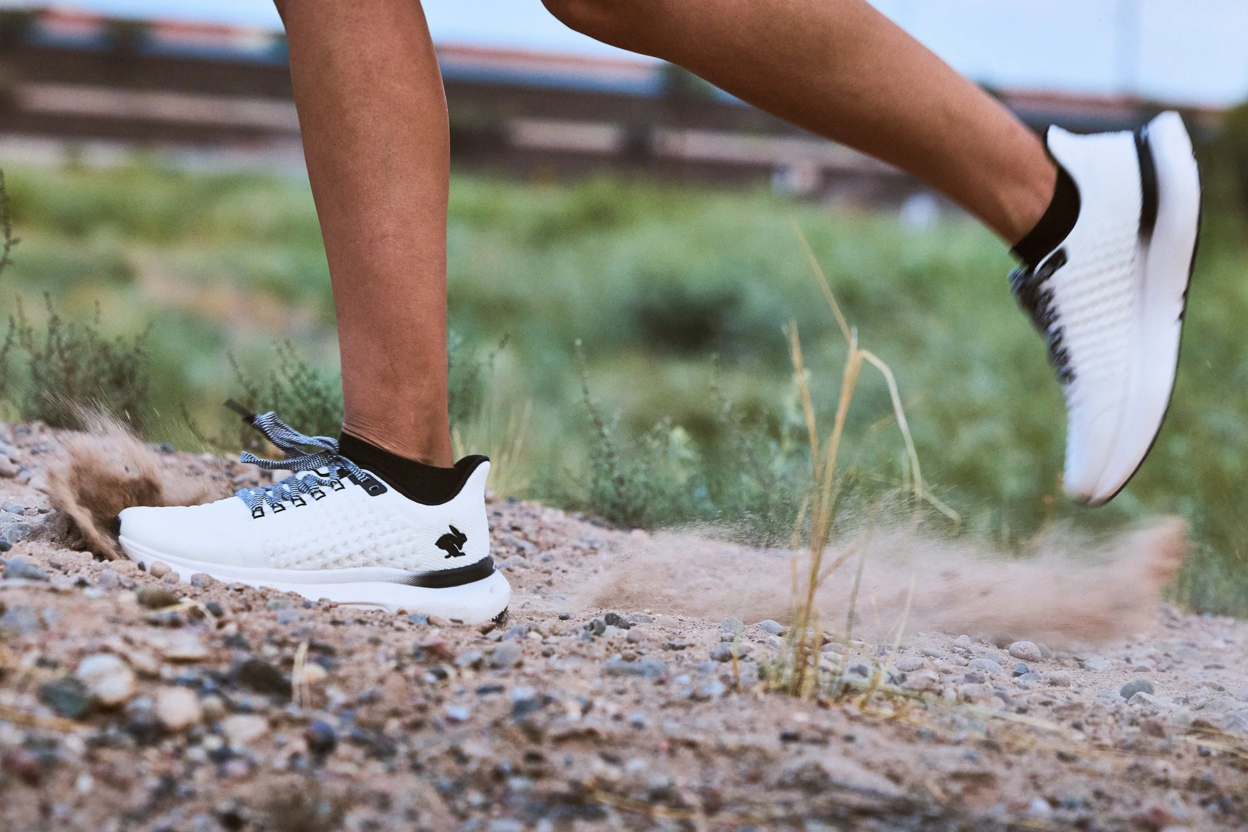 Close-up of a person's legs and feet wearing white athletic shoes with black accents, running or walking on a rocky dirt trail with greenery in the background.