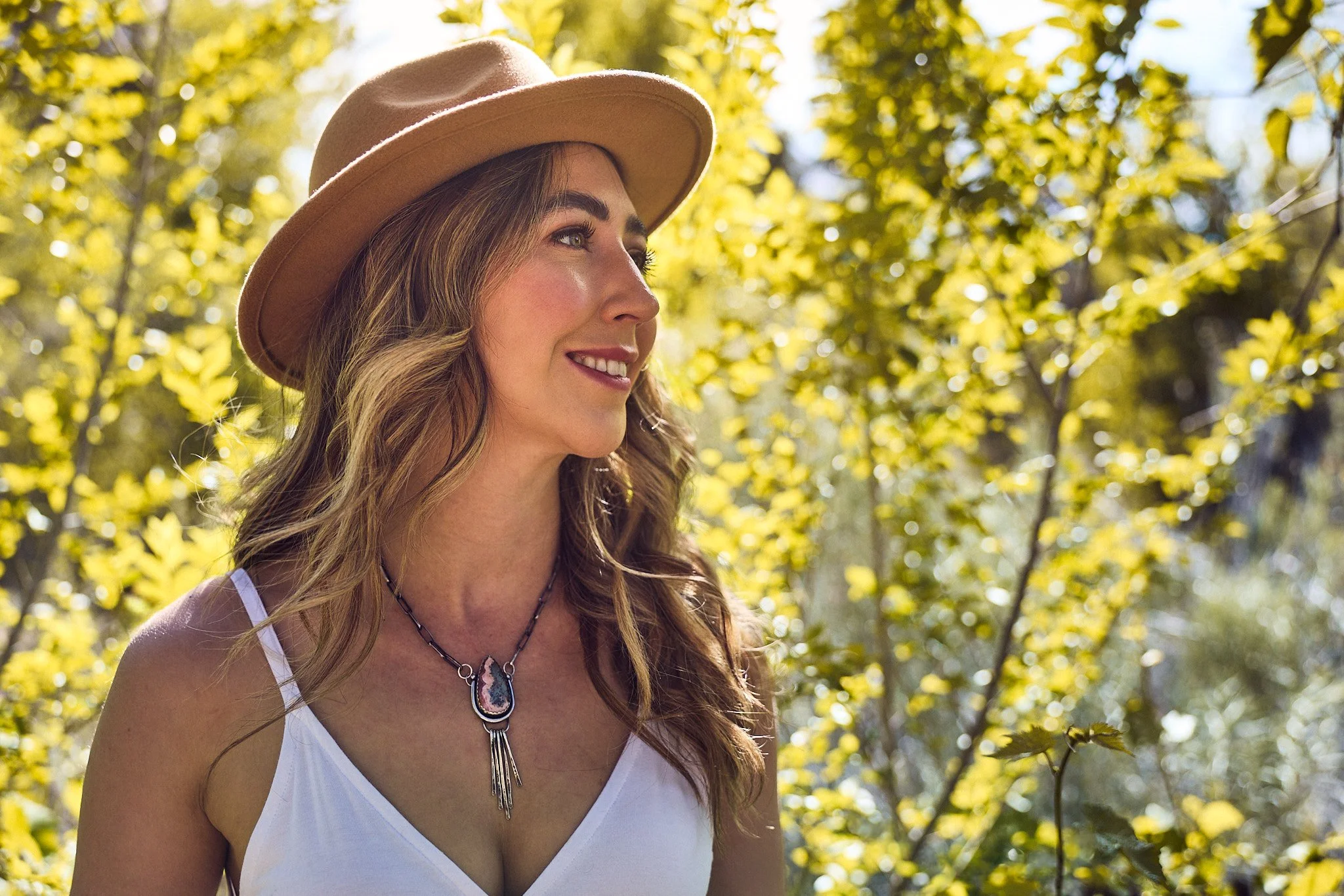A young woman with long, wavy hair wearing a beige hat and a white tank top, standing outdoors among yellow foliage, smiling and looking to the side, with a necklace featuring a pendant.