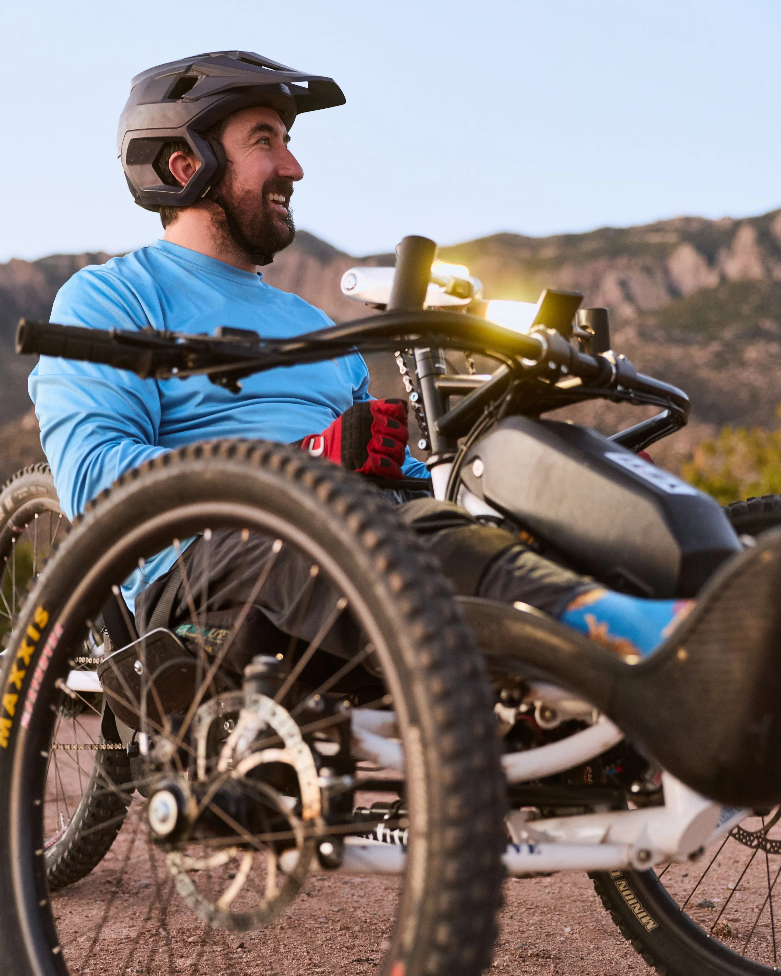 A smiling man sitting on a mountain biking trail wearing a helmet and gloves, with mountains visible in the background.