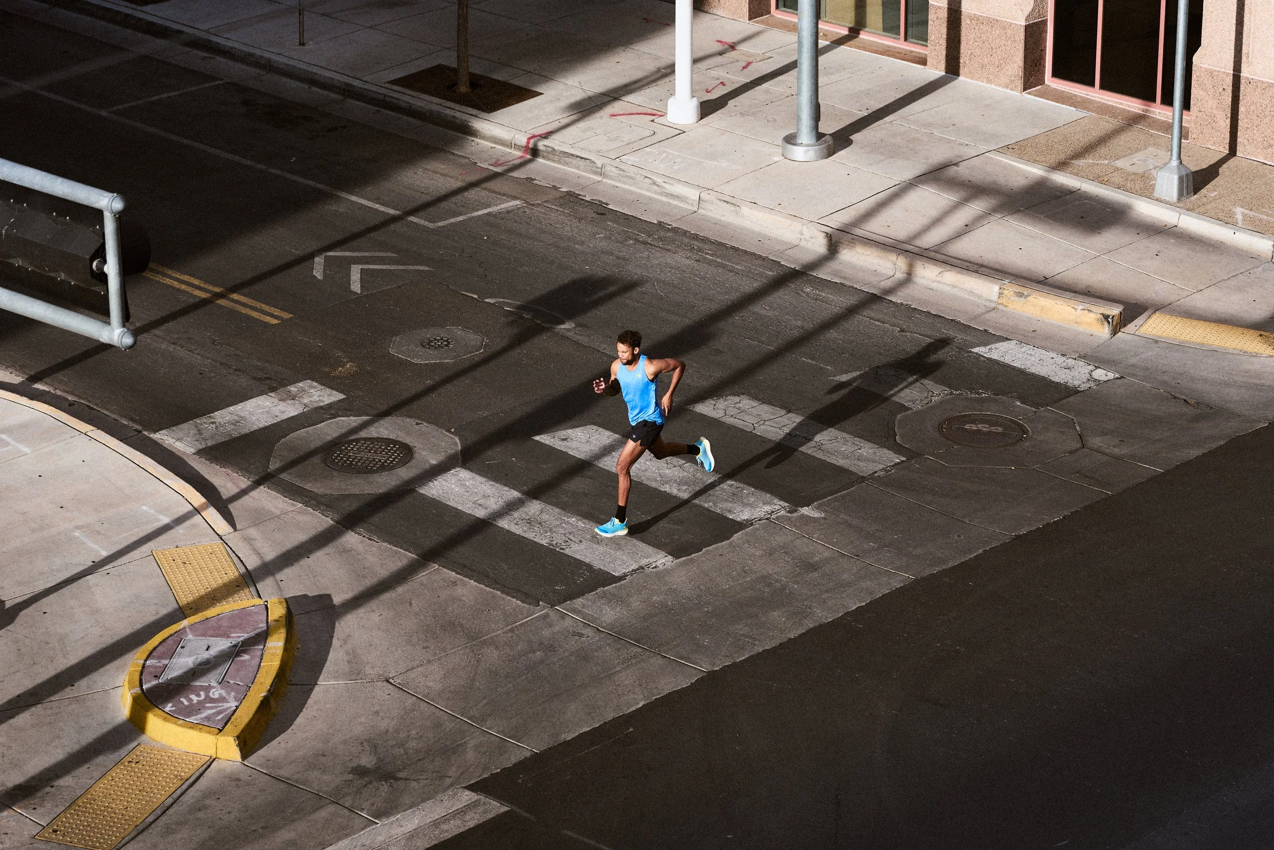 A man jogging on a city street at night, wearing a blue tank top, black shorts, and blue running shoes. The street is mostly empty, with some street markings and urban infrastructure visible.
