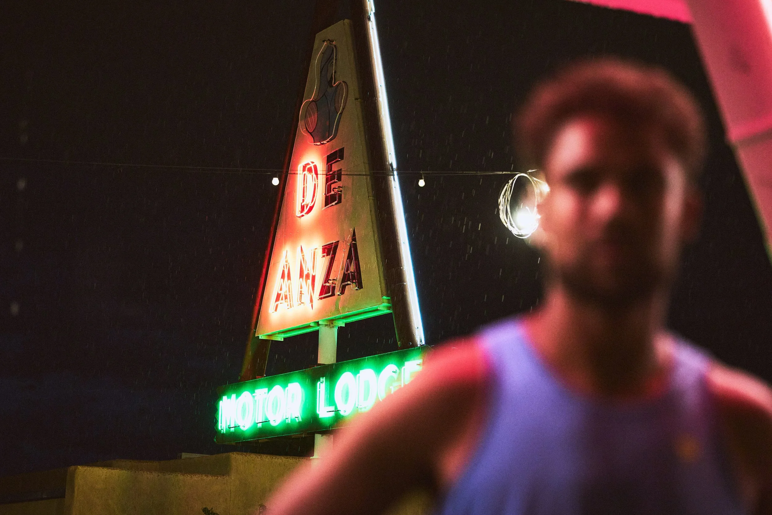 Nighttime view of a neon sign for a motor lodge, illuminated in red and green colors, with a person partially visible in the foreground and raindrops falling around.