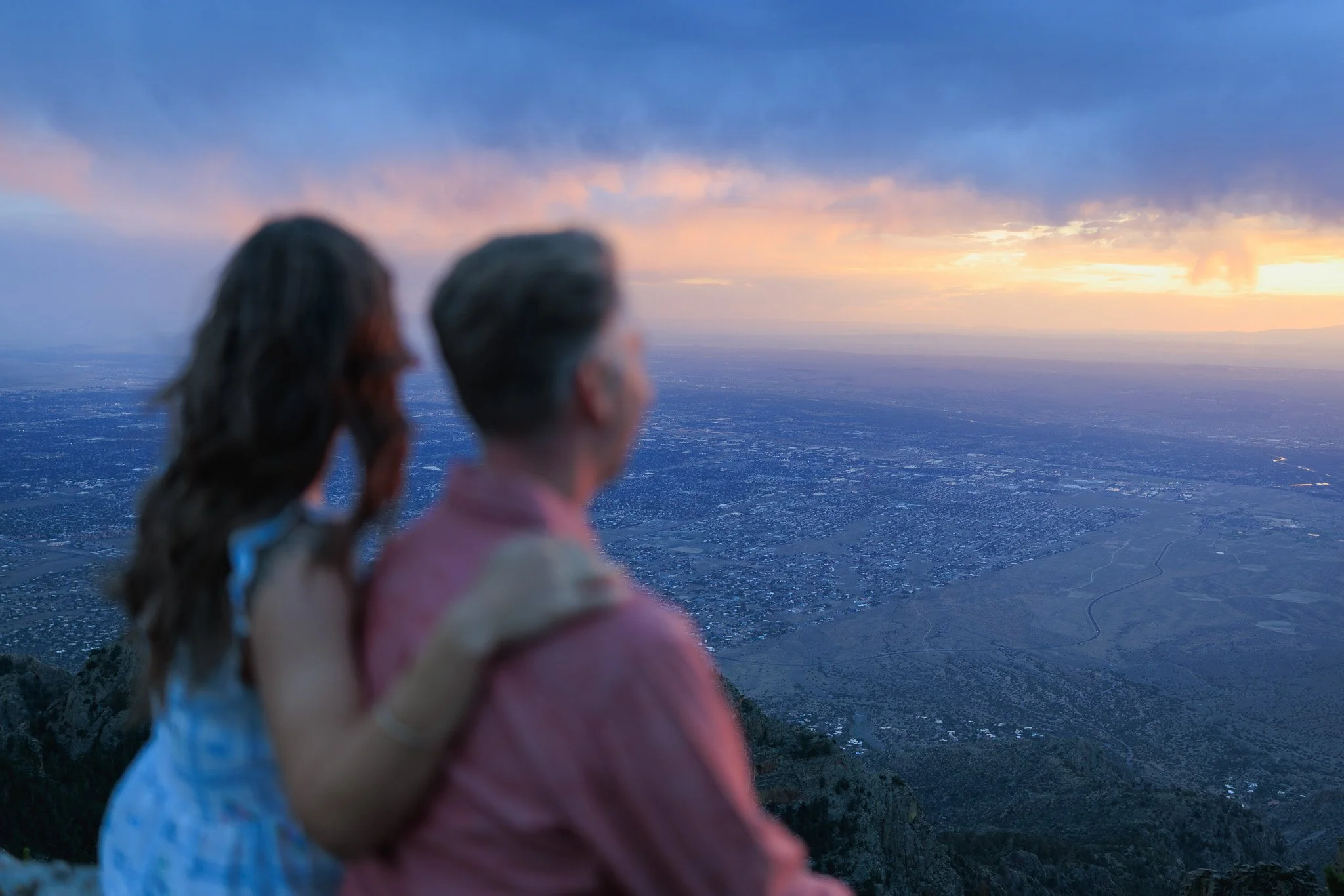 Two people, a girl with a ponytail and a woman with short gray hair, holding hands and looking out over the landscape during sunset from a mountain or hilltop.