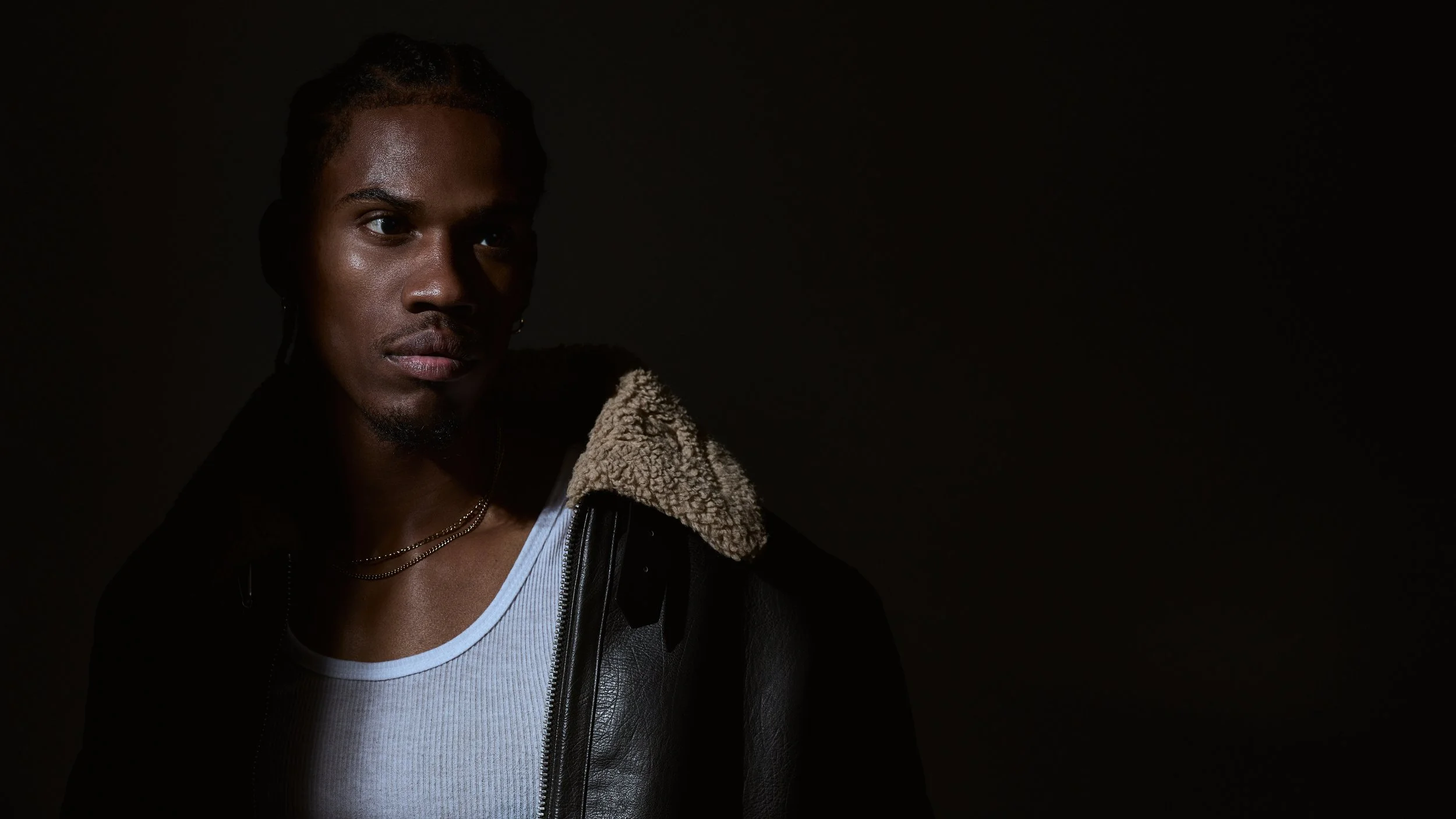 Portrait of a young woman with dark skin and braided hair, wearing a white tank top, black leather jacket with a shearling collar, and layered necklaces, against a dark background.