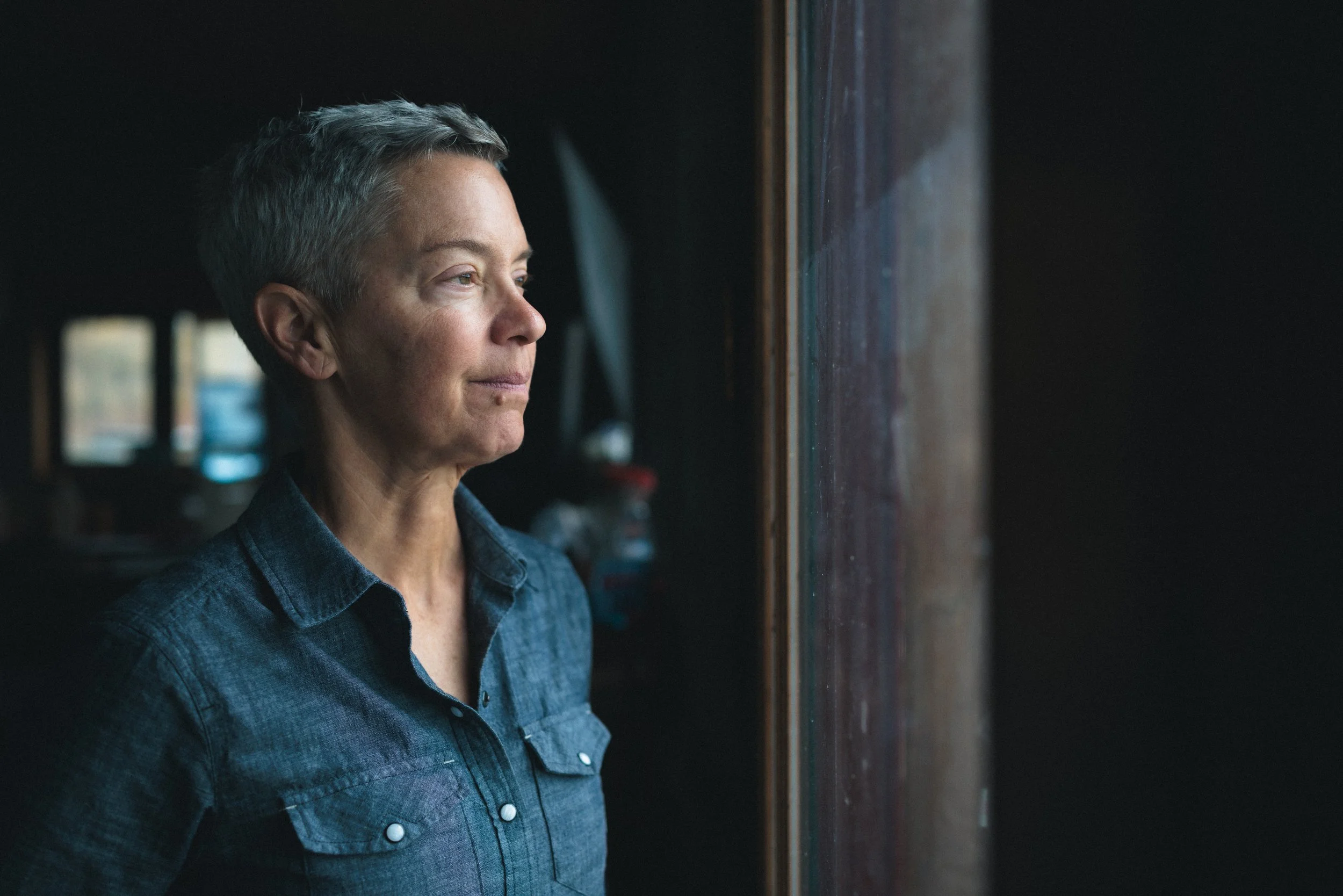 Woman with short gray hair looking thoughtfully out a window in a dimly lit room.