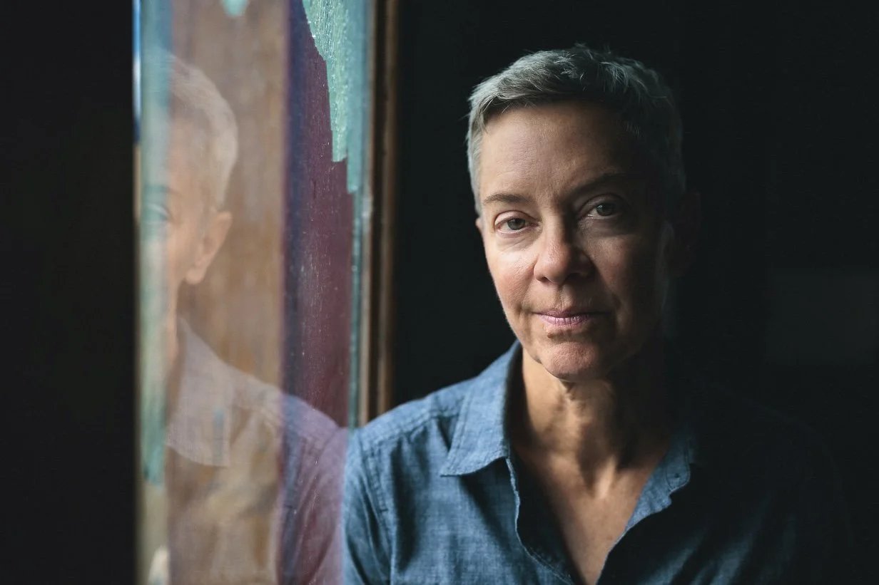 A middle-aged woman with short gray hair looking out a window, reflected in the glass, wearing a blue shirt.