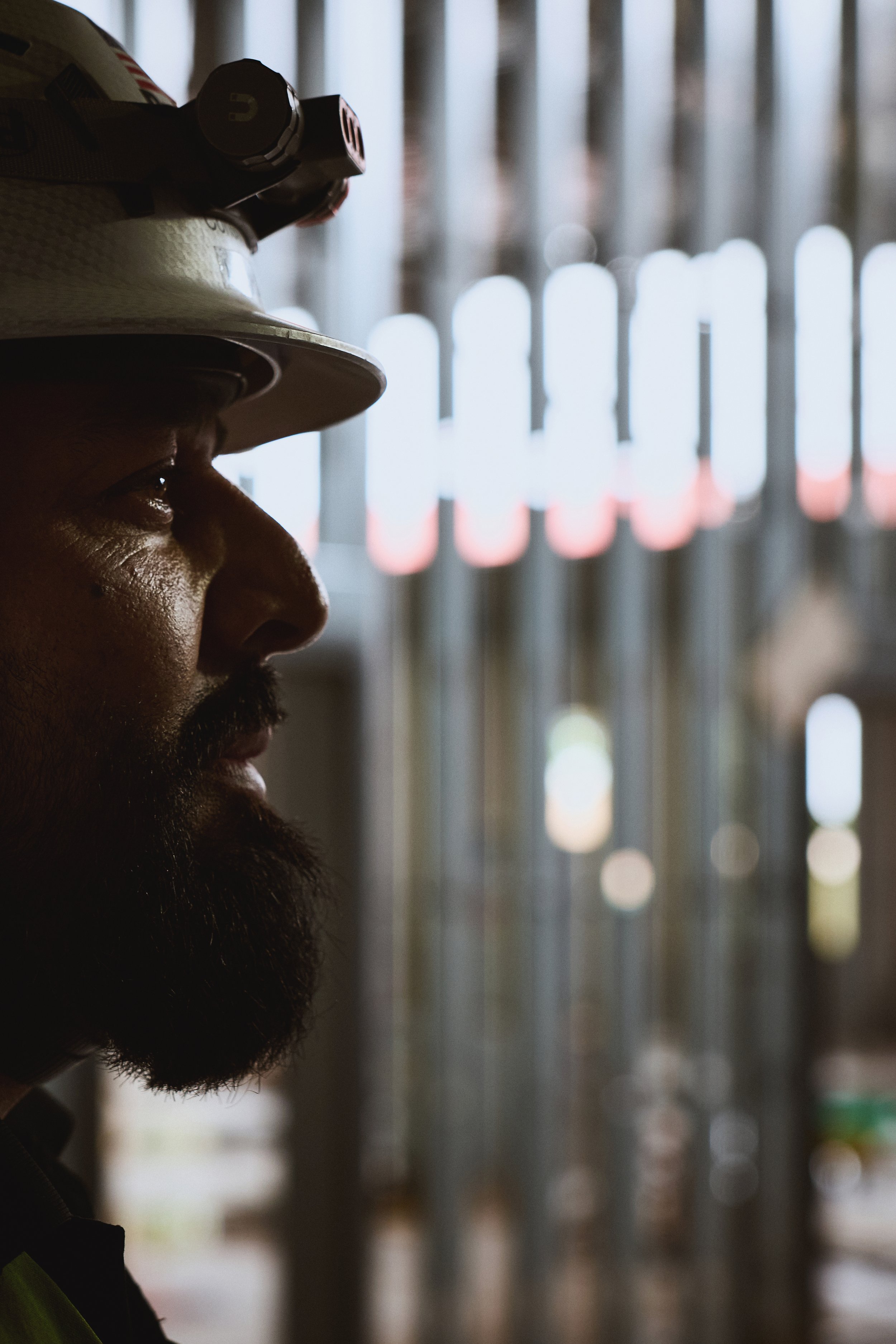 A man wearing a white safety helmet and a headlamp, seen in profile, standing in a dimly lit environment with blurred vertical pipe or railing structures in the background.