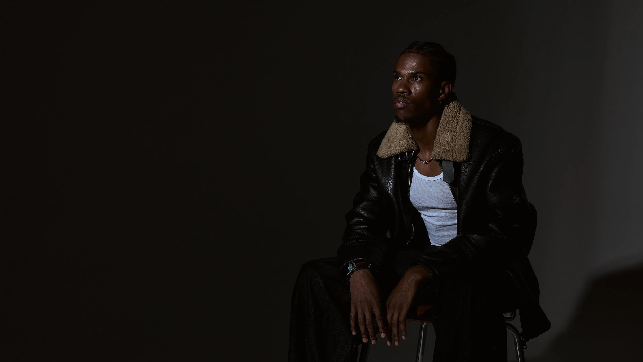 A young man with dreadlocks and earrings sits on a chair against a dark background, wearing a black leather jacket with a shearling collar and a white shirt.