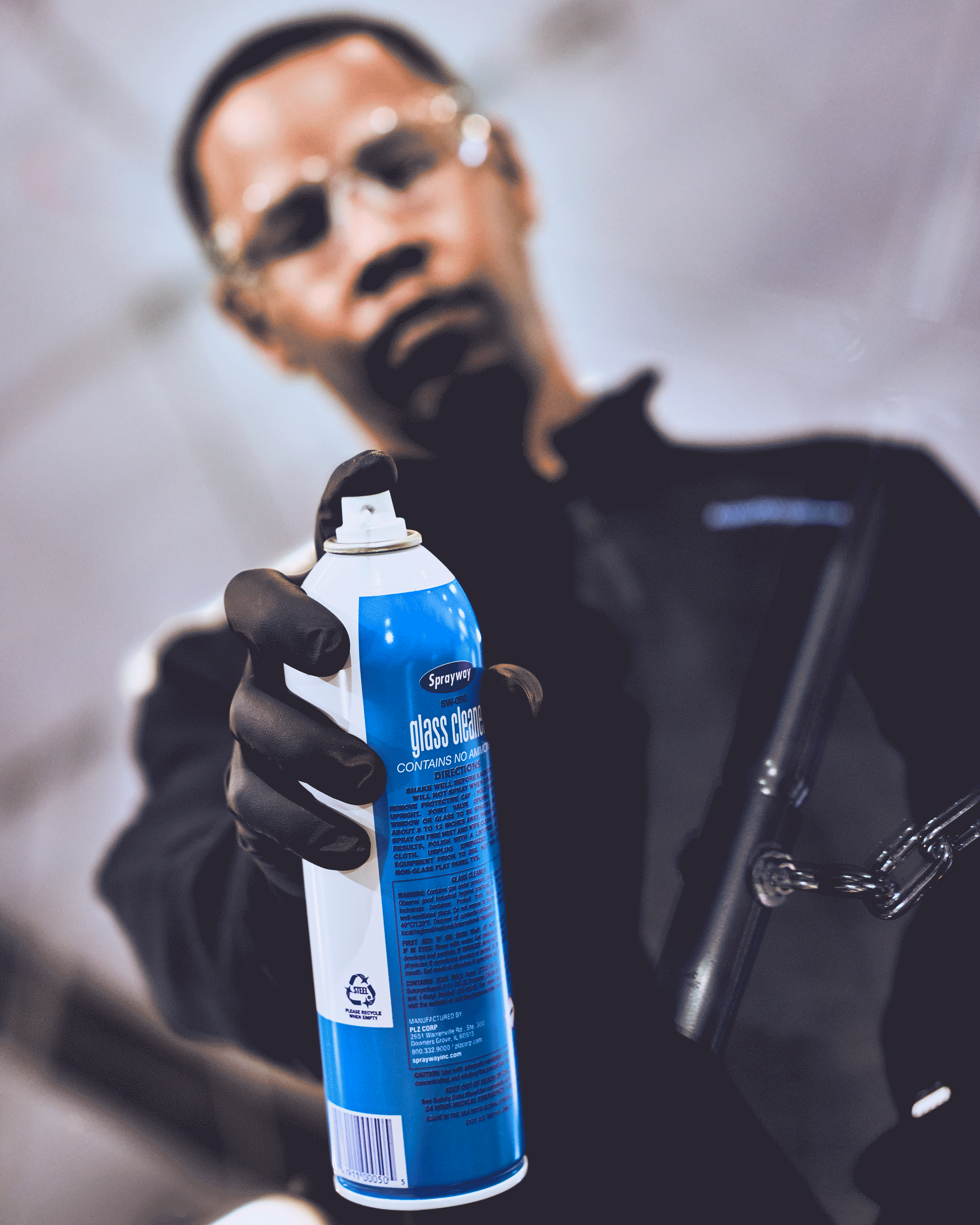 A person wearing black gloves holding a can of spray glass cleaner, with the person's face blurry in the background.