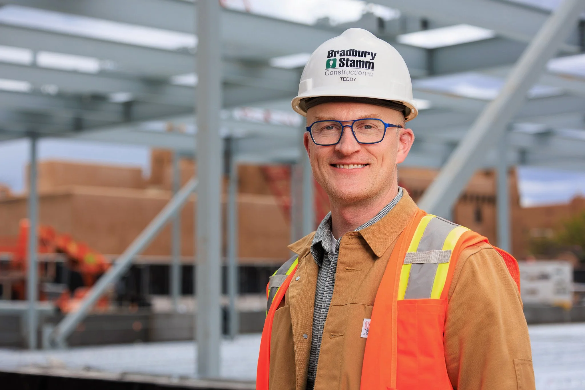 A smiling construction worker wearing glasses, a white hard hat with "Bradbury Stamm Construction" and "TEDDY" written on it, and a high-visibility safety vest, standing in front of a construction site with metal framework.