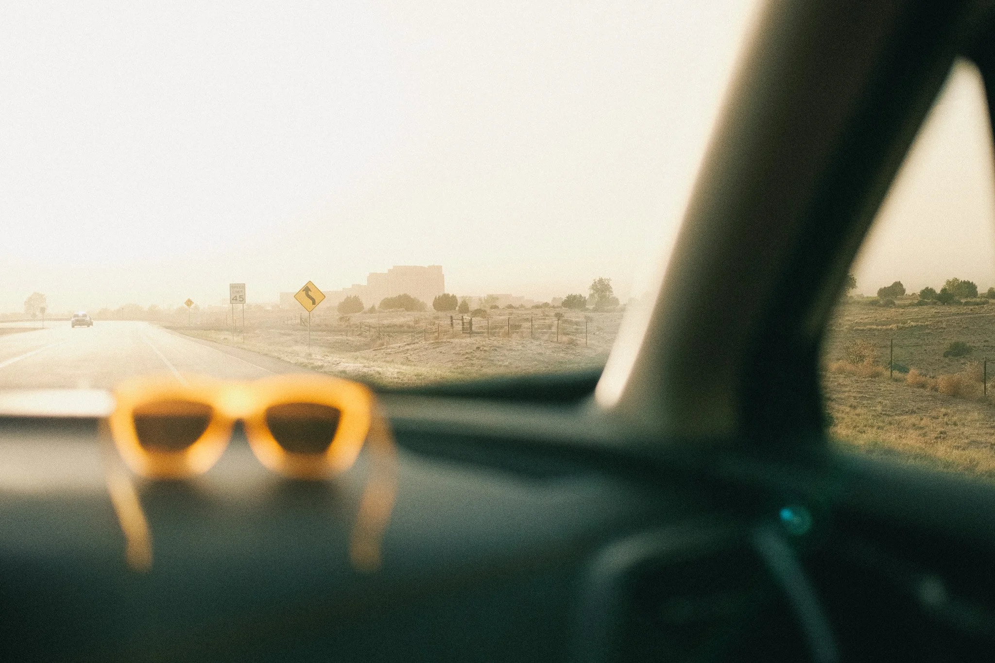 View from inside a car looking out the windshield at a highway with a car ahead, hazy weather, road signs including a curve warning, and distant buildings and trees on a rural landscape.