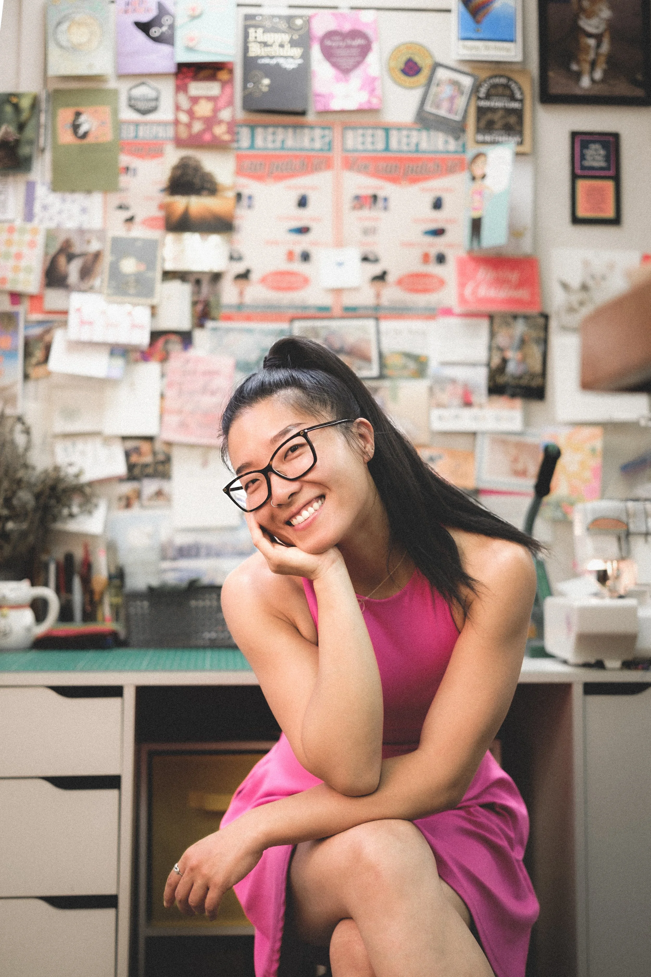 A woman with long dark hair, glasses, and a bright smile, wearing a pink dress, sitting in a cluttered workspace with a wall covered in handmade cards, photos, and drawings.