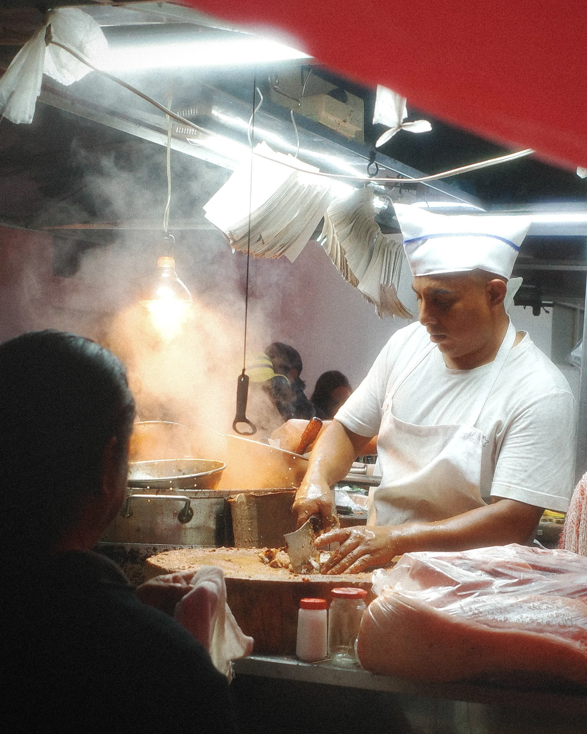 A chef cooking in a busy restaurant kitchen with steam rising, wearing a white chef hat and uniform, preparing food at a large cooking station.