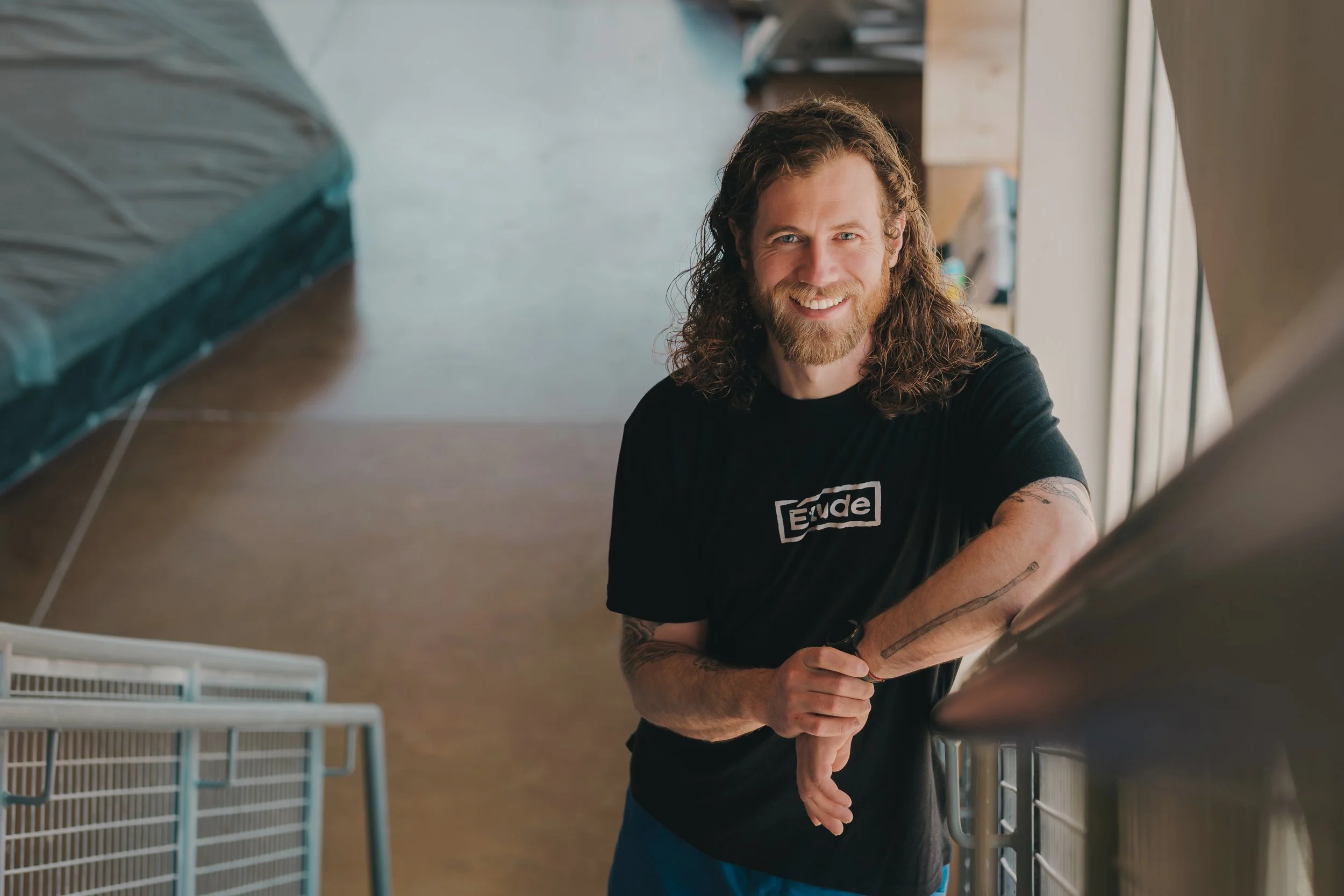 A smiling man with long curly hair and a beard, wearing a black T-shirt, is leaning on a metal pet crate inside a room with large windows, tan flooring, and household items in the background.