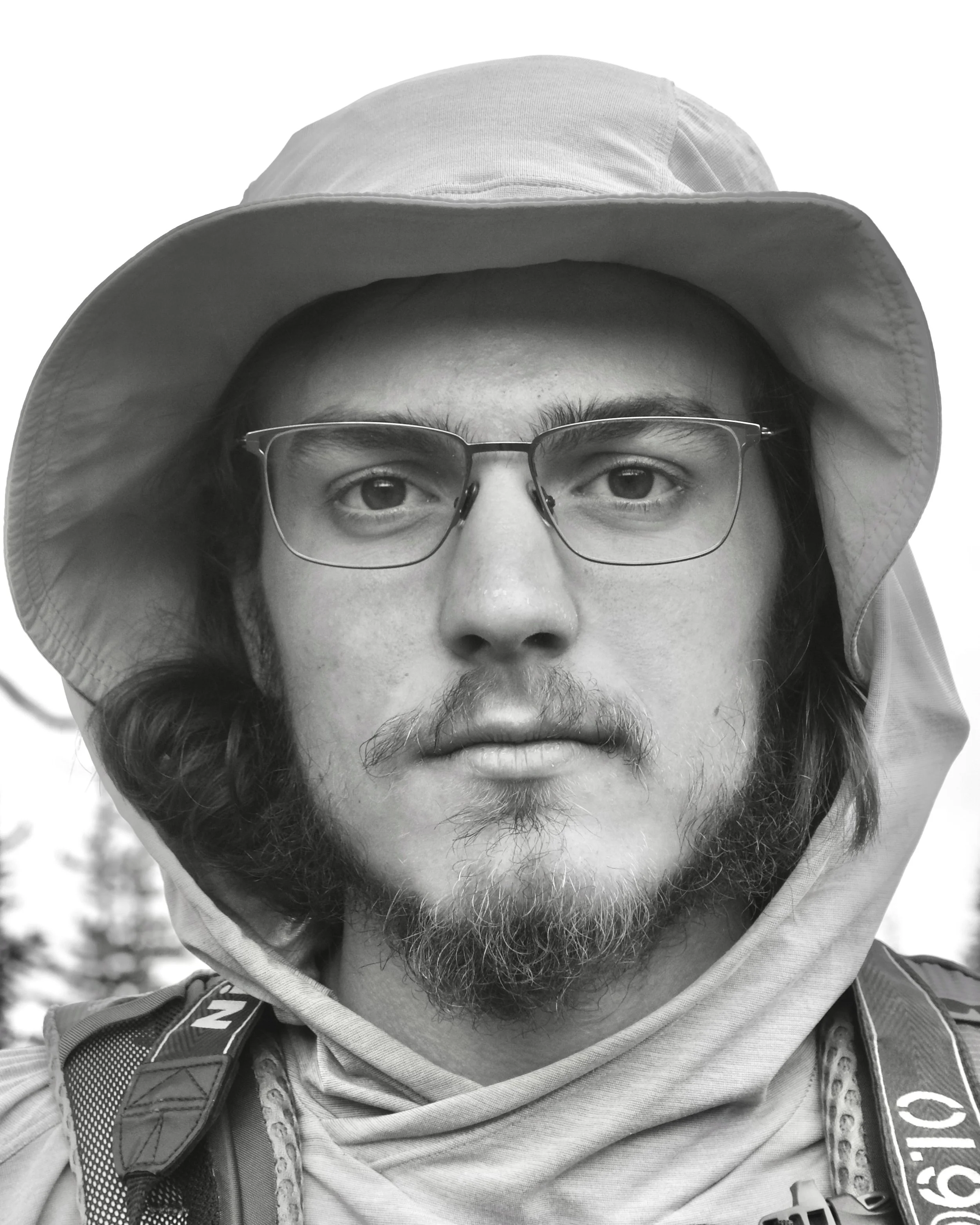 Close-up of a person's face, wearing glasses, a wide-brimmed hat, and a hoodie, with a beard and mustache, in black and white.
