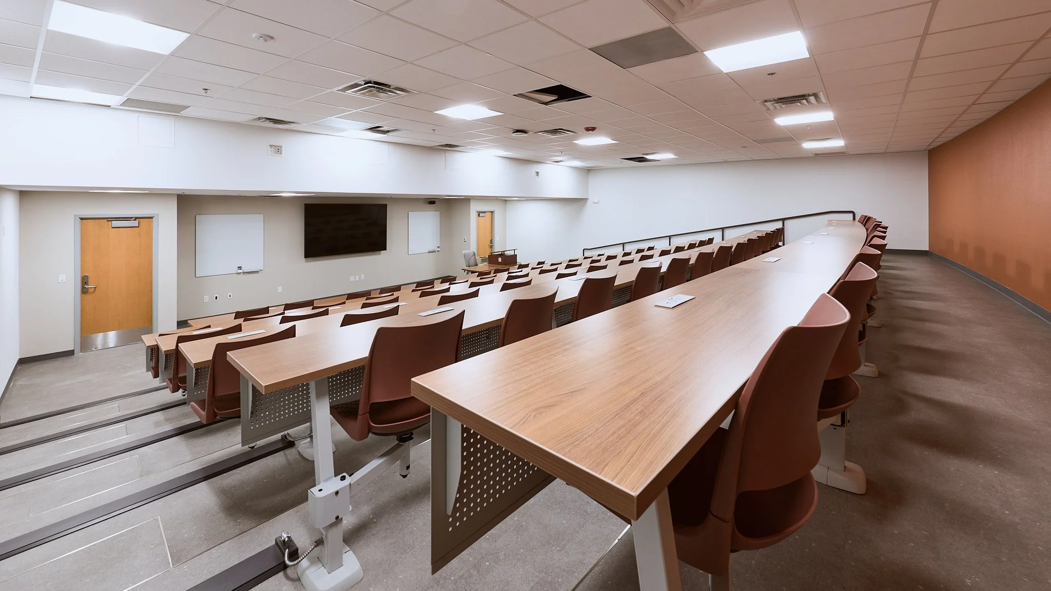 Empty university classroom with long curved table, brown chairs, white walls, ceiling lights, and a large black screen at the front.