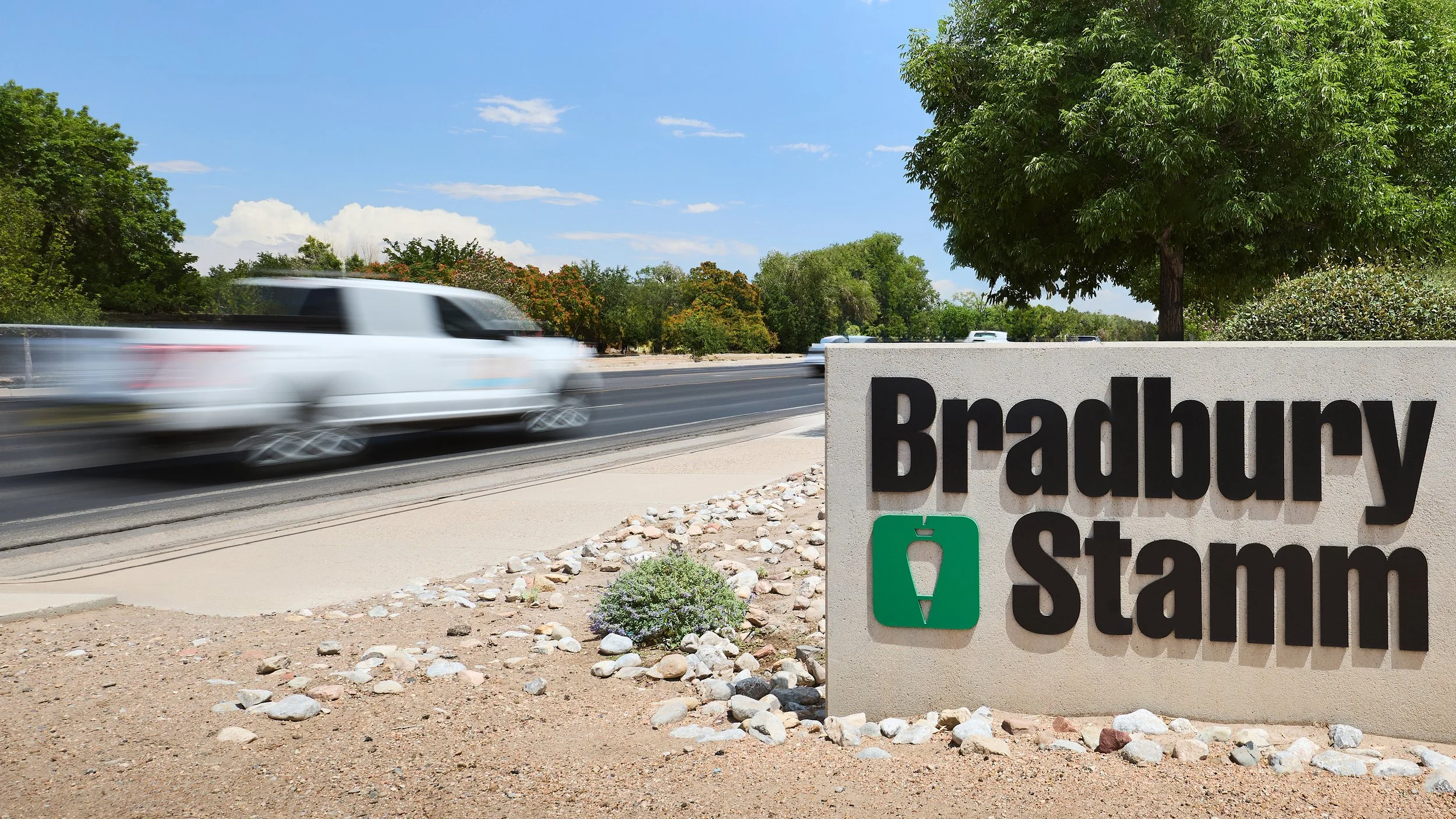 Speeding cars on a highway near a large sign that reads 'Brauery Stamn' with a green water fountain icon, trees and a blue sky in the background.