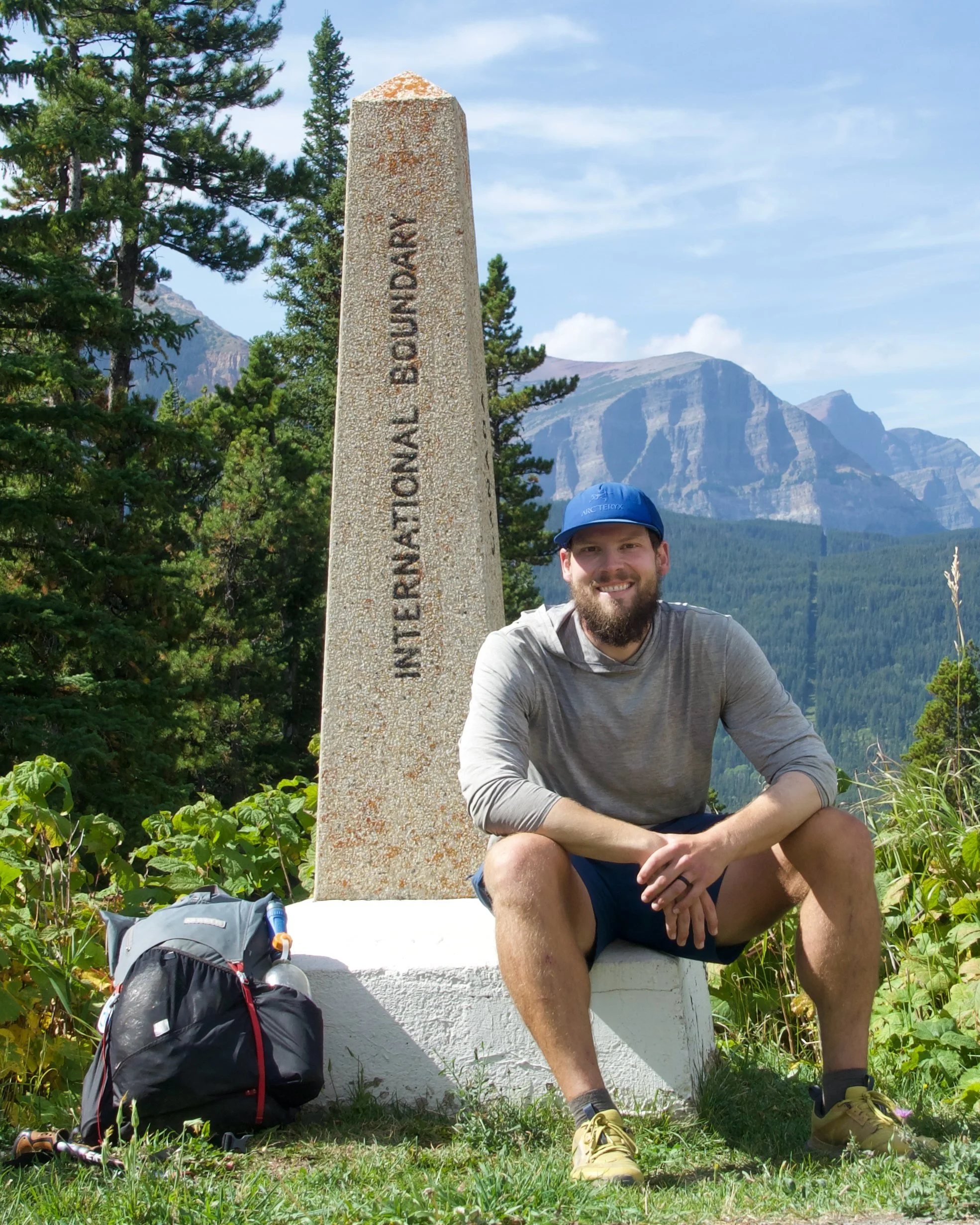 A man with a beard and gray shirt sitting on a white monument with a mountain and trees in the background. A backpack and hiking gear are on the ground near him.