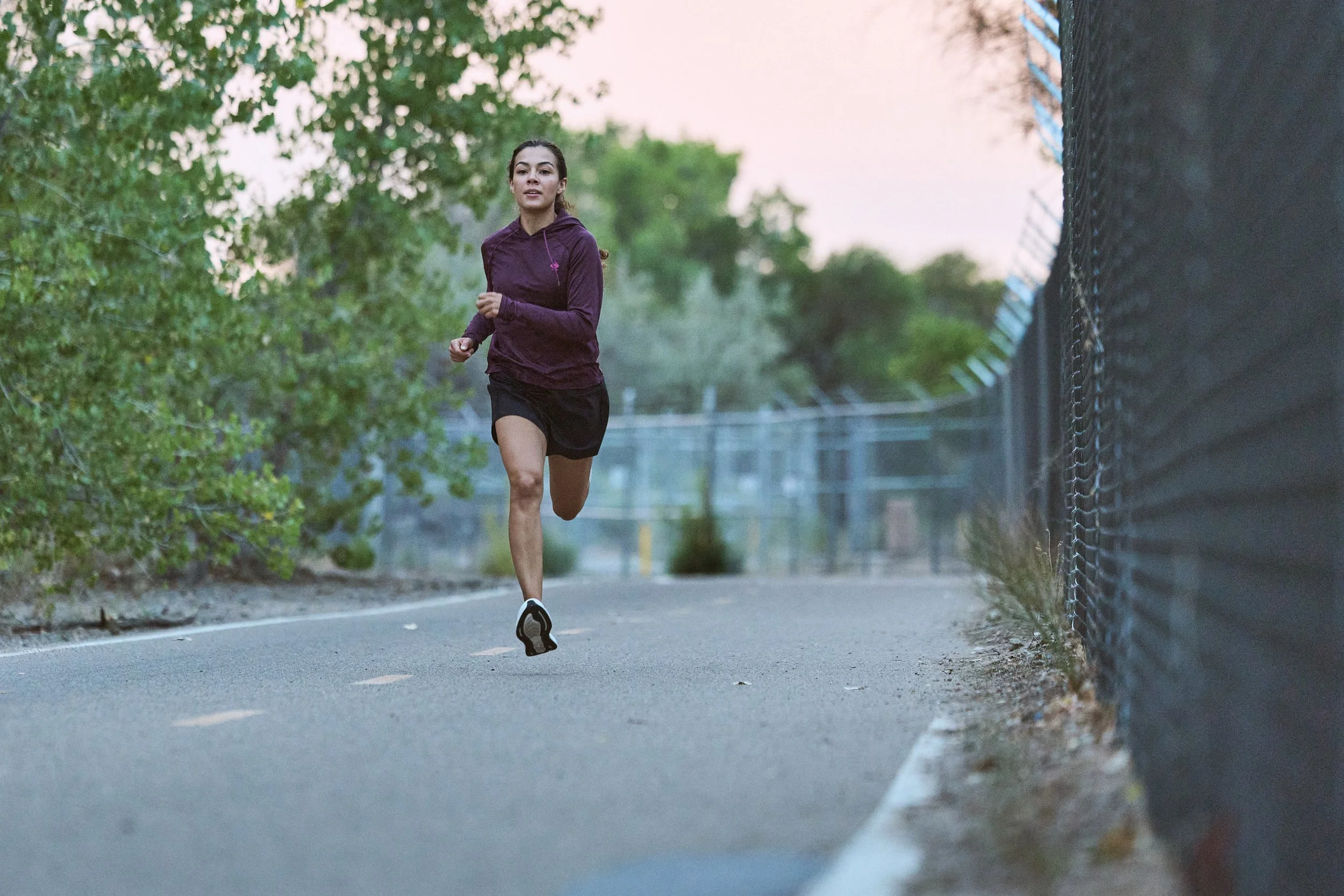 A woman jogging on a paved trail near a chain-link fence with trees and a pale sky in the background during dawn or dusk.