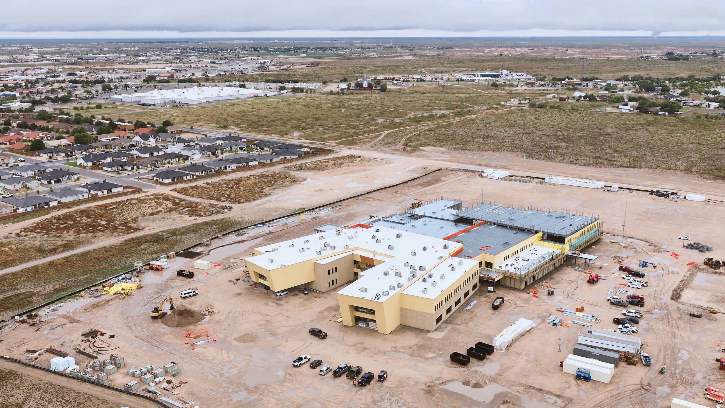 Aerial view of a construction site with a yellow building, construction equipment, and vehicles, adjacent to a residential area with rows of houses, and a distant commercial or industrial area under an overcast sky.