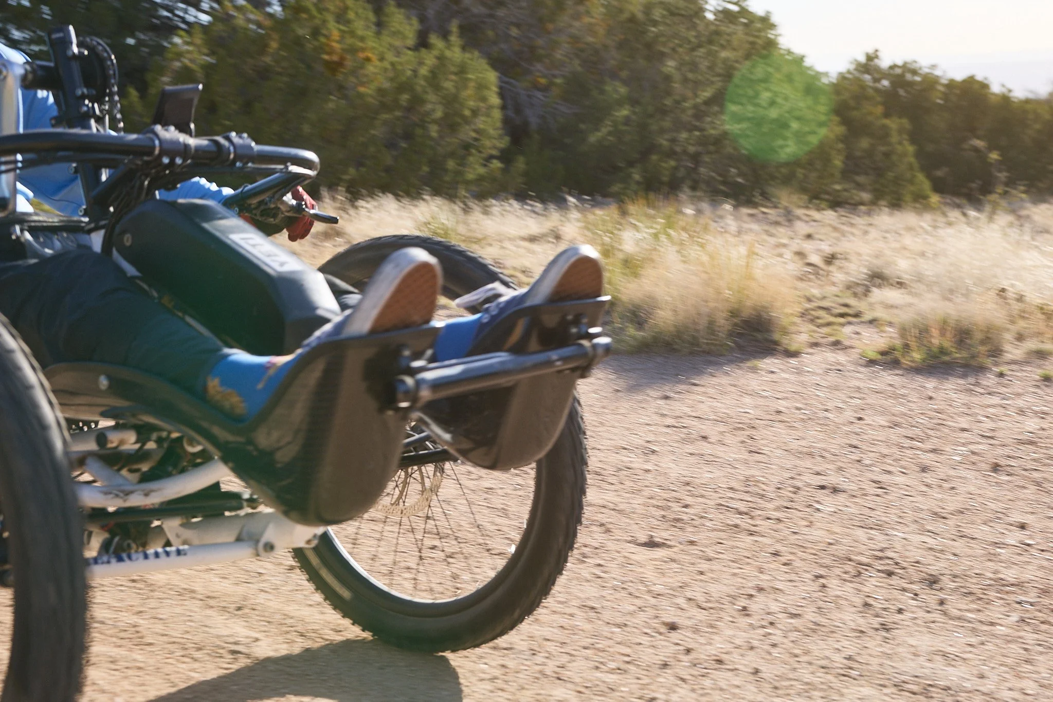 A person lying in a mountain bike with their feet up, riding on a dirt trail through a forested area with green trees and dry grass.