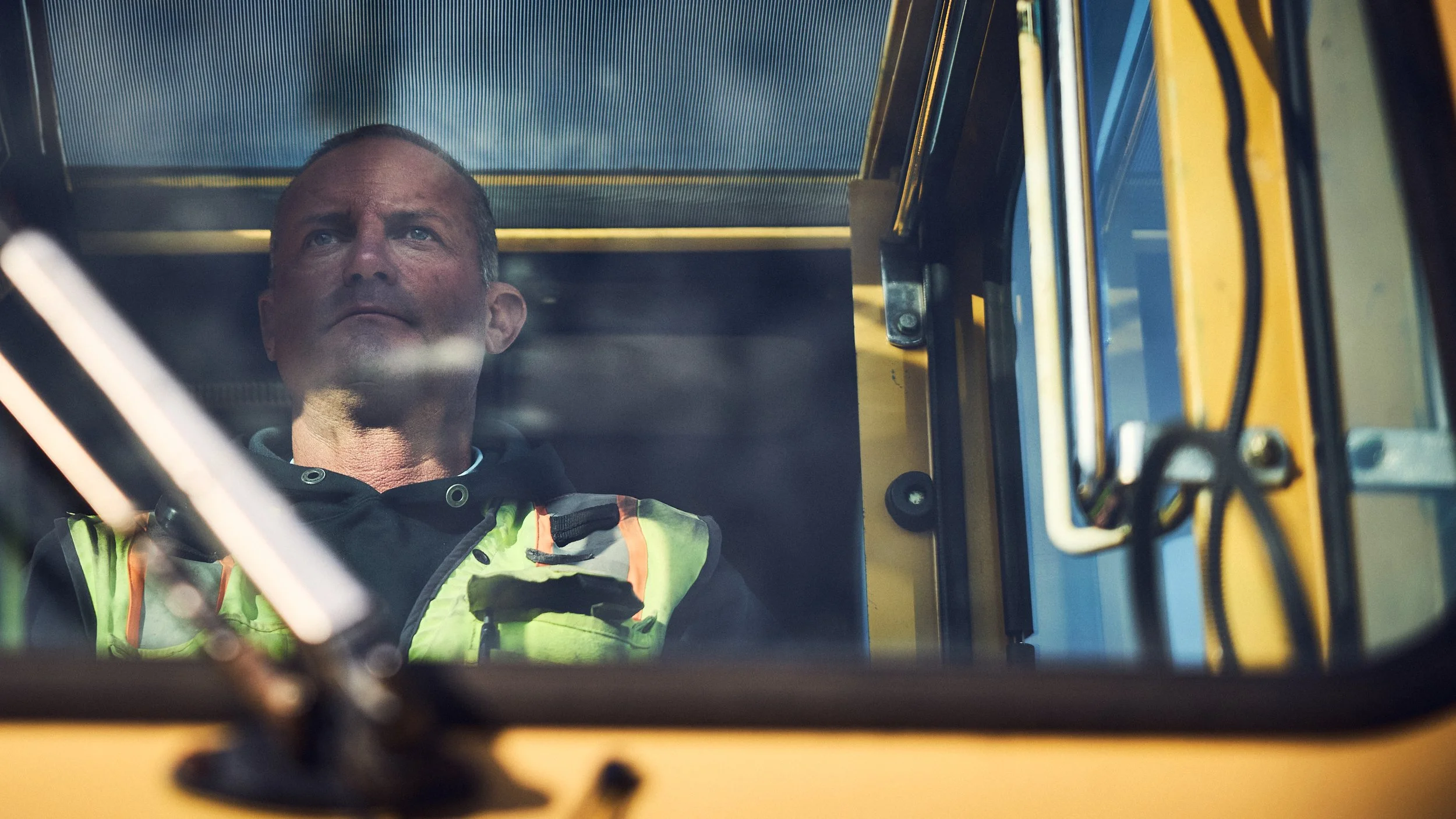 A man in a black jacket with a neon yellow safety vest sits inside a yellow construction vehicle, visible through the window.