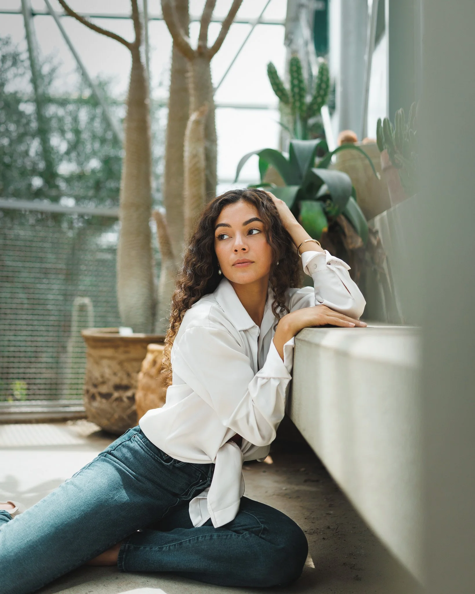 A woman with curly hair, wearing a white shirt and jeans, is sitting on the floor and leaning against a white surface, with her hand resting on her head. She is in a greenhouse or indoor plant area with various plants and trees in the background.