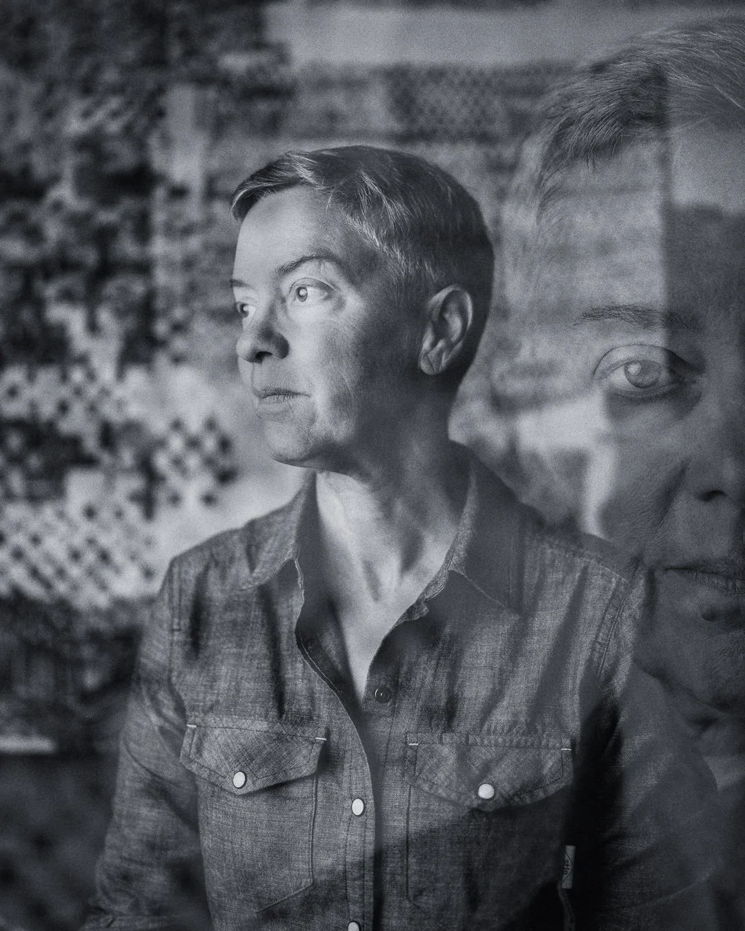 A black-and-white photo of a woman looking to the side with a reflection of her face overlapping her shoulder.