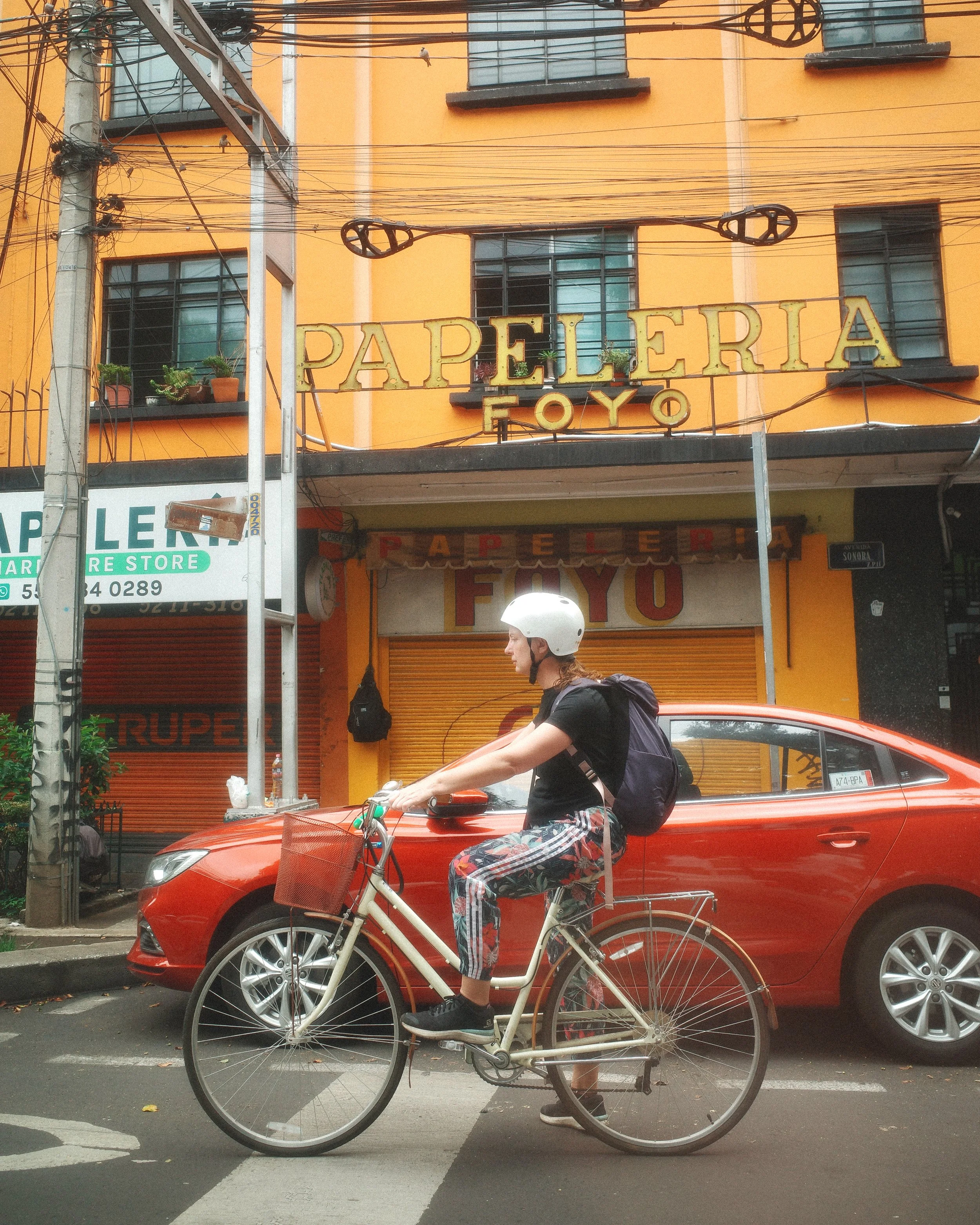 A person riding a white bicycle on the street with a red car parked behind them, in front of a yellow building with sign that reads 'Papeleria Foyo' and black window frames. The person is wearing a white helmet, black shirt, patterned pants, and carr