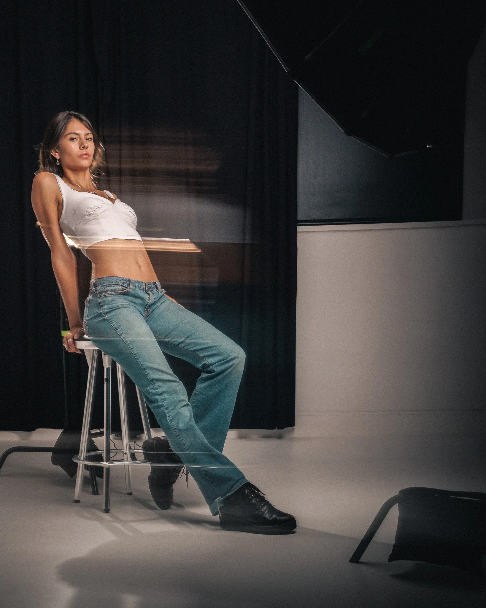 A young woman with dark hair, wearing a white crop top and blue jeans, sitting on a stool against a dark background, with artistic motion blur effects around her.