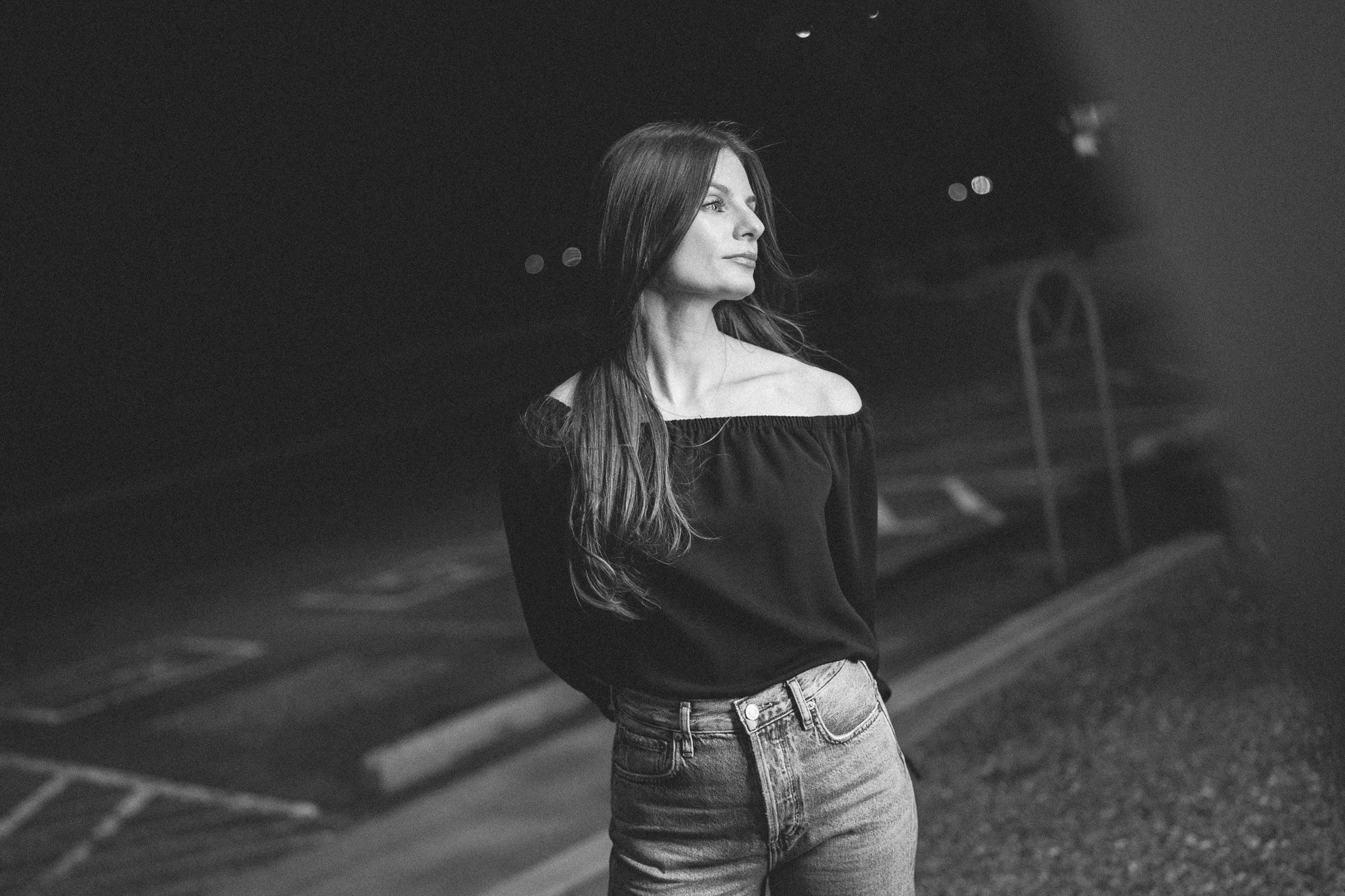 A black-and-white photo of a young woman with long hair standing in an outdoor parking lot at night, looking to her right, wearing an off-the-shoulder top and jeans.