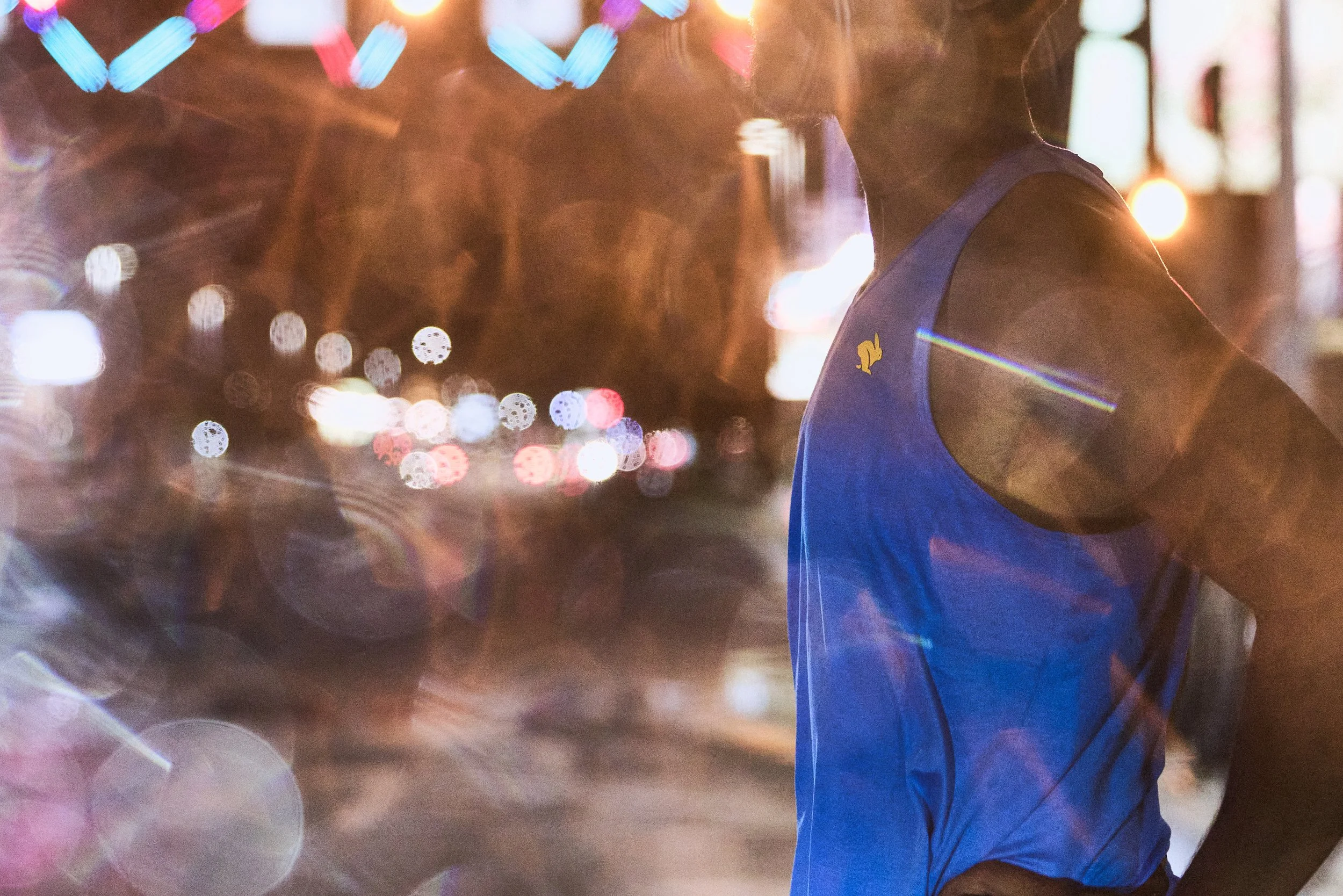 A person in a blue shirt, seen from the side, standing outdoors at night with city streetlights and blurred colorful lights in the background.