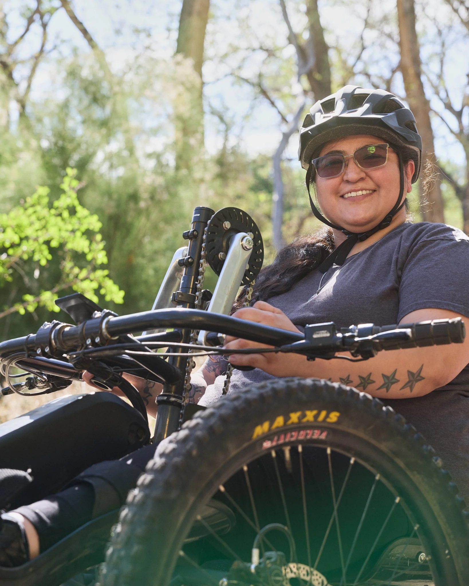 A woman wearing glasses, a black helmet, and a gray t-shirt is smiling while sitting on a mountain bike outdoors in a green, wooded area.