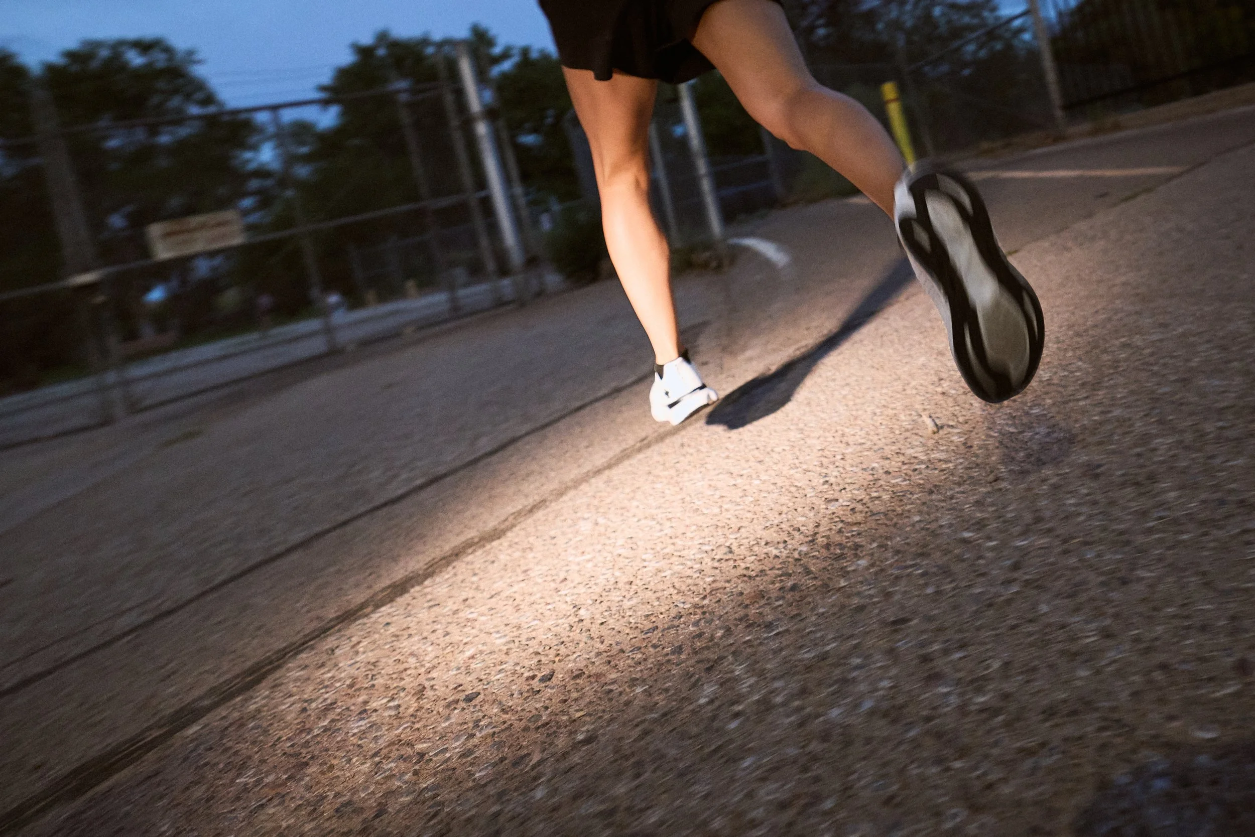 A person wearing white sneakers and black shorts running on a paved path during dusk or dawn.