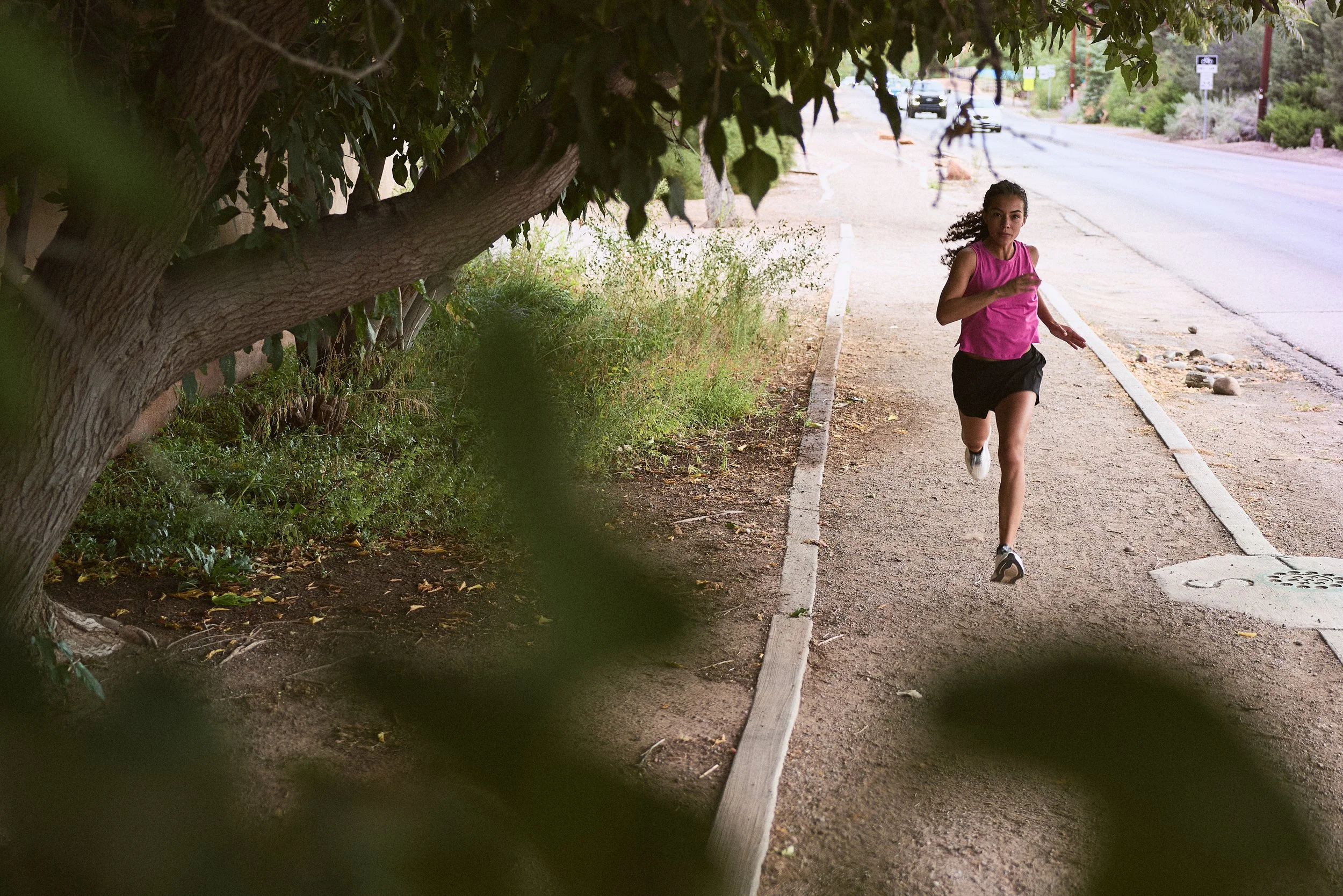 A young woman running on a sidewalk next to a tree with green leaves, wearing a pink tank top and black shorts, in a suburban area with cars and houses in the background.
