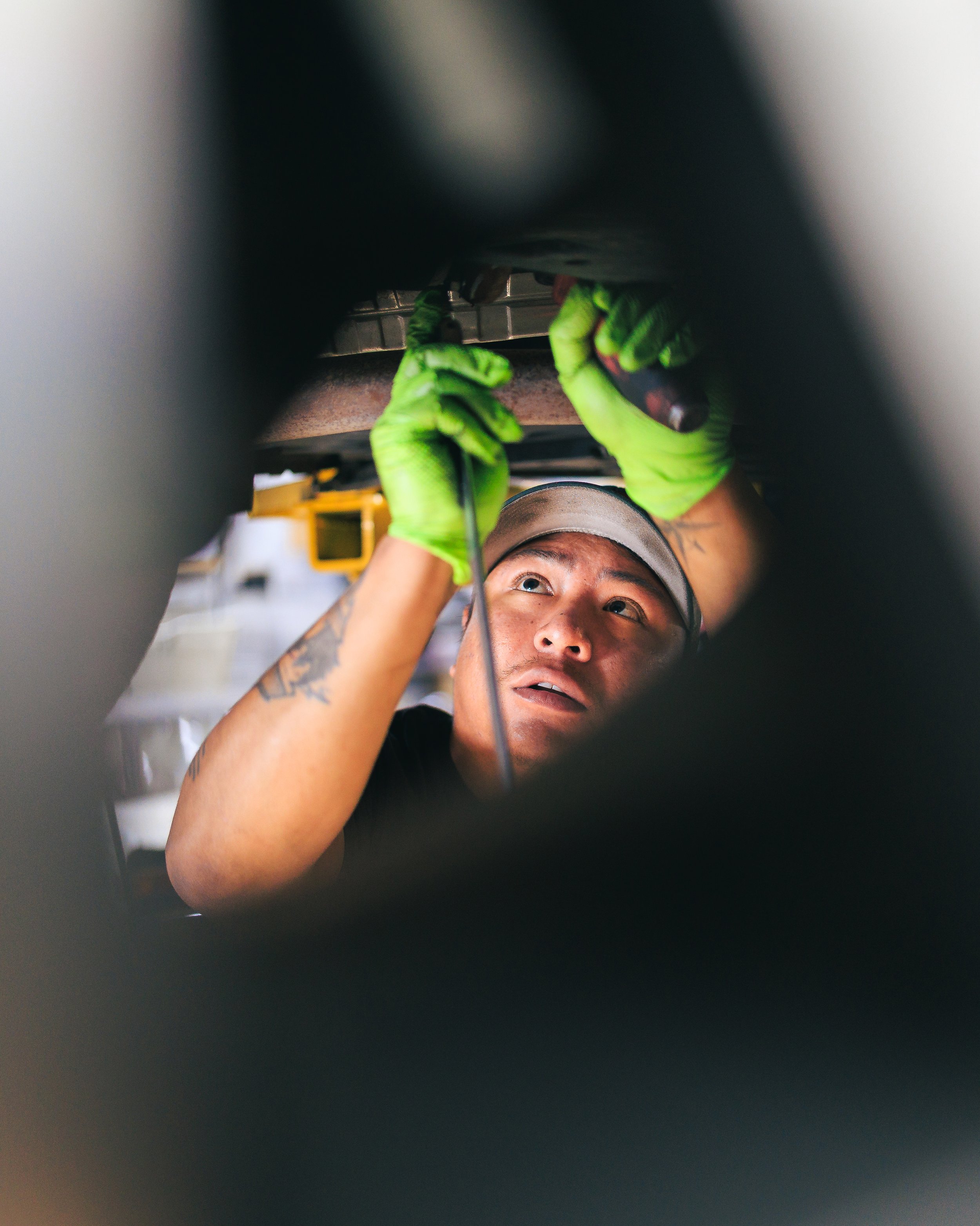 A person wearing green gloves working underneath a washing machine or dishwasher, possibly repairing or inspecting it.