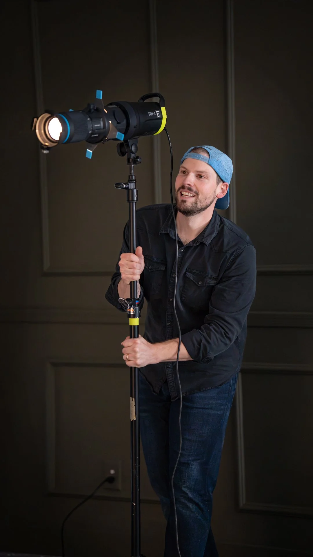 A man wearing a blue cap backwards and a black shirt is adjusting a professional camera mounted on a tripod in a dark room.