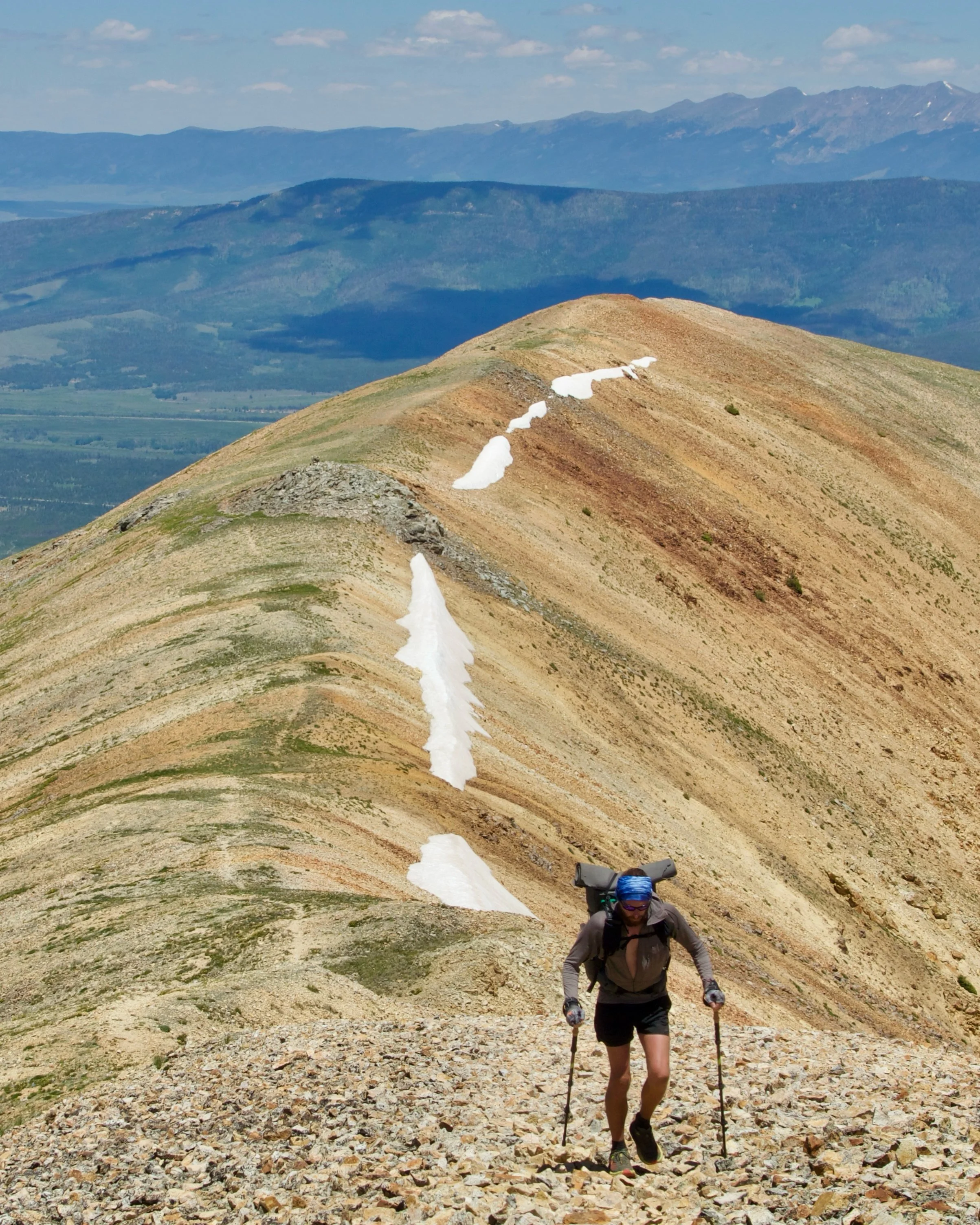 A person hiking on a rocky trail on a mountain with patches of snow, with a mountain range in the background under a partly cloudy sky.