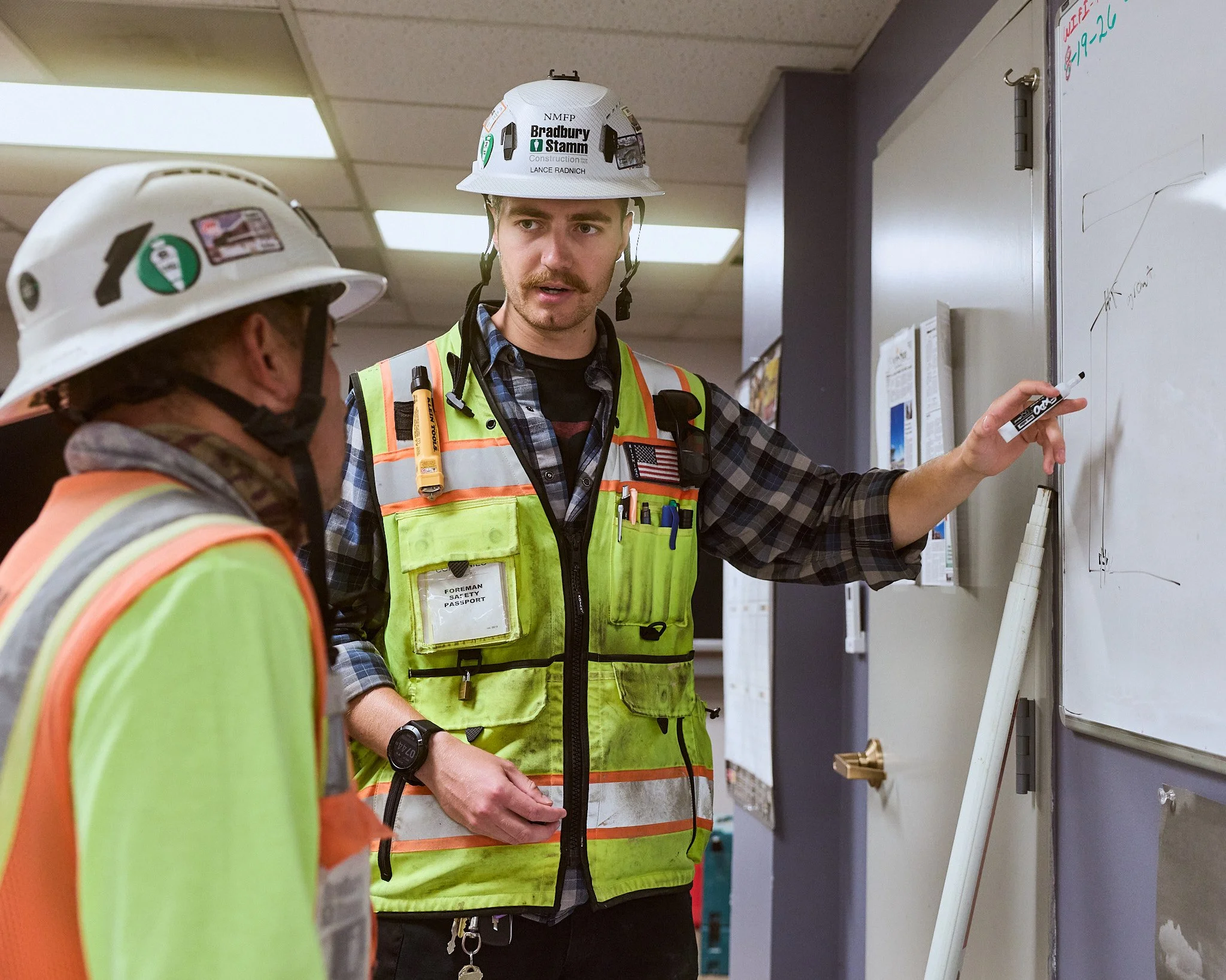 Two construction workers wearing safety helmets and reflective vests in a conversation in front of a whiteboard in an indoor setting.