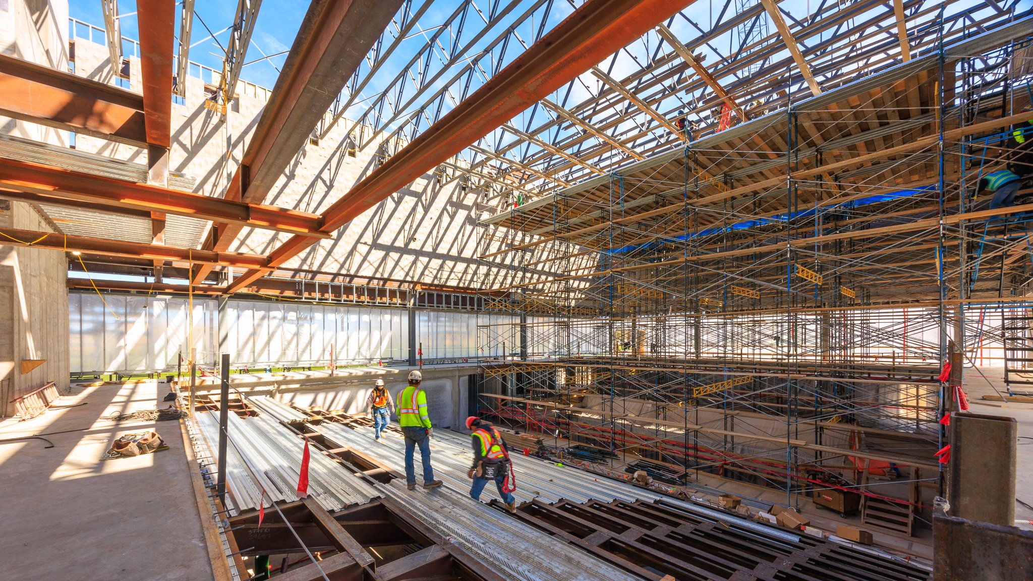 Construction workers in safety vests and helmets working on a large building under construction, with extensive scaffolding, steel beams, and a partially built roof structure.