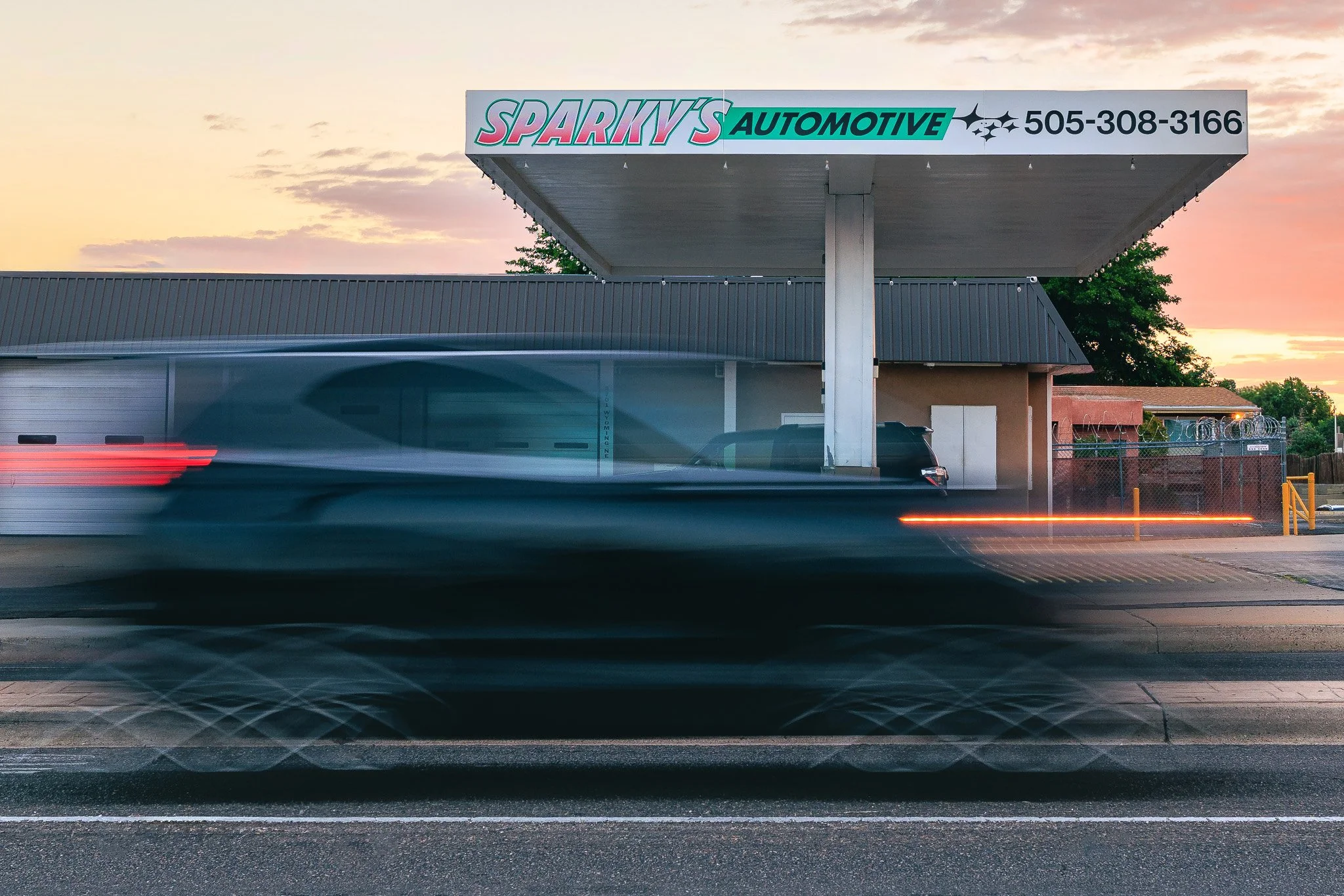Sign for Sparky's Automotive shop at sunset, with a free-standing canopy covering part of the parking area. A car passes by, creating light trails, and the sky has pink and orange hues with some clouds.