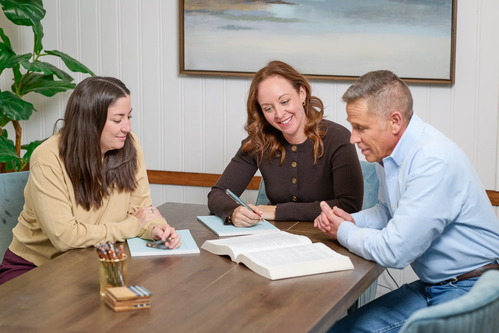 Four people sitting around a wooden table in a room with white paneling walls, engaged in a discussion or meeting. One woman is smiling and taking notes, with other participants listening attentively.