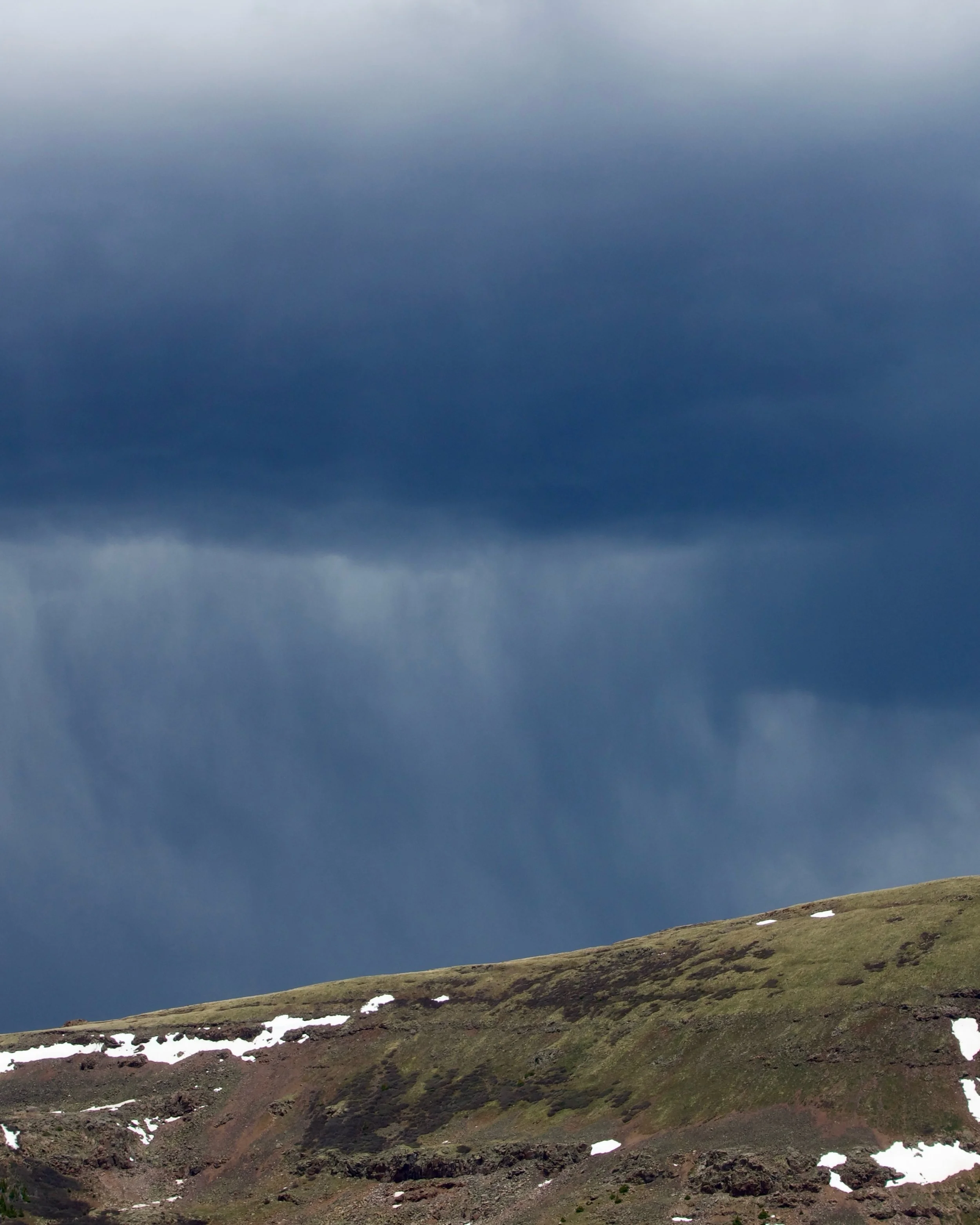 View of a mountain landscape with a grassy slope, patches of snow, dark rocky terrain, and a cloudy sky overhead.