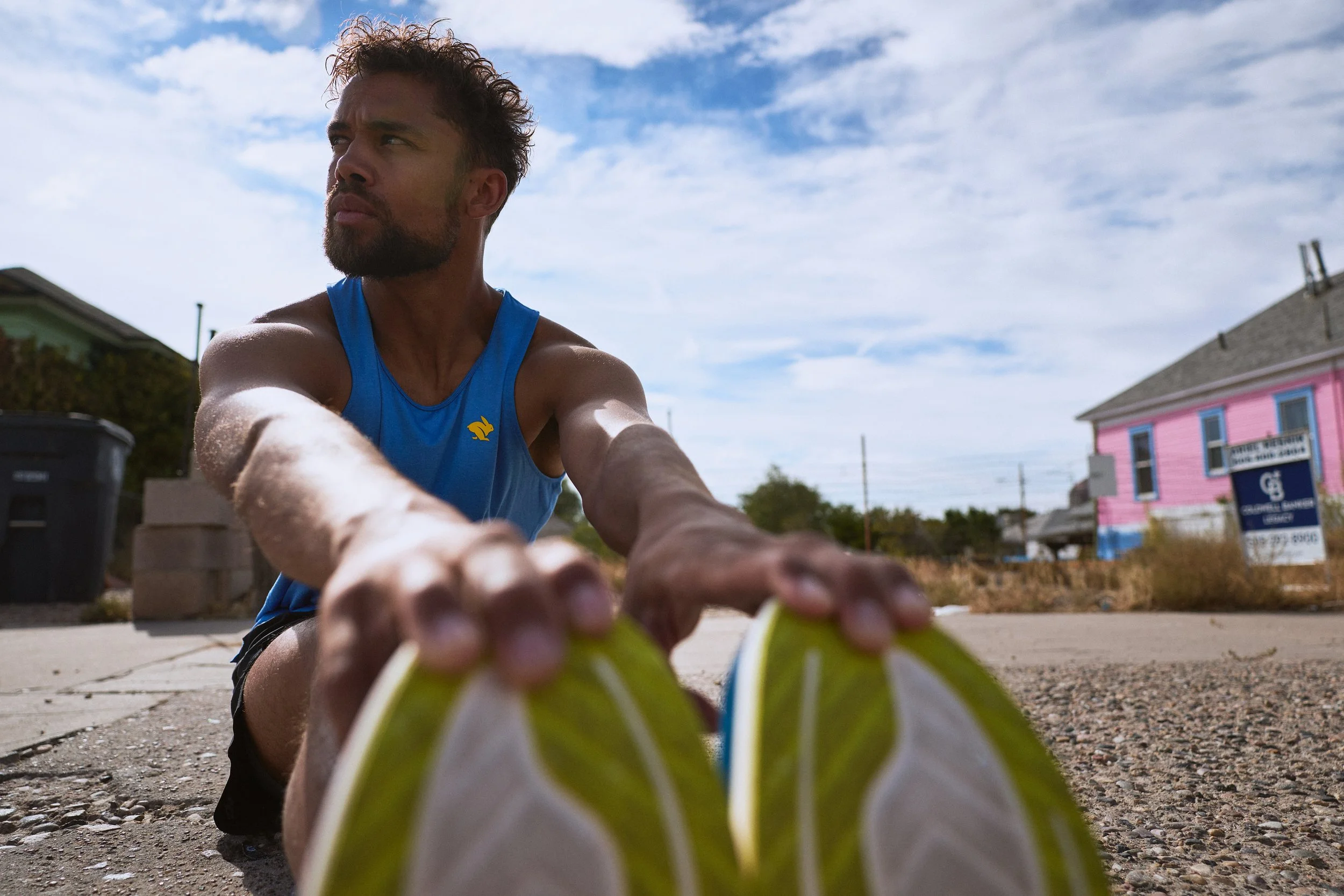 Man stretching on the sidewalk during a sunny day, wearing a blue athletic tank top, with a colorful building in the background.